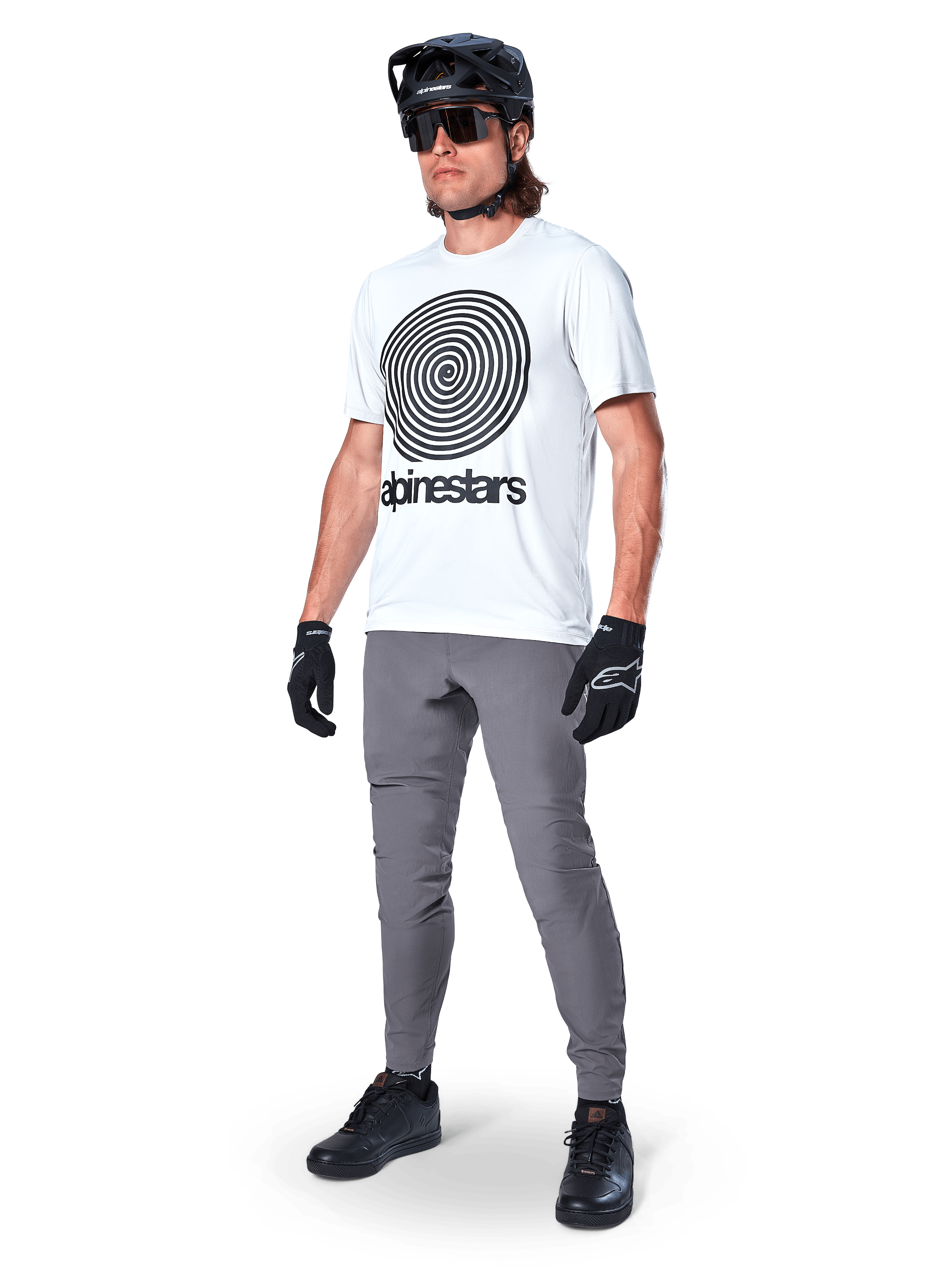 Alpinestars A-Dura Byxor, MTB-byxor, mörkgrå, bärs med vit t-shirt med spiralgrafik, svart MTB-hjälm, solglasögon och svarta handskar, slitstarkt 2-vägs stretchtyg för terrängcykling