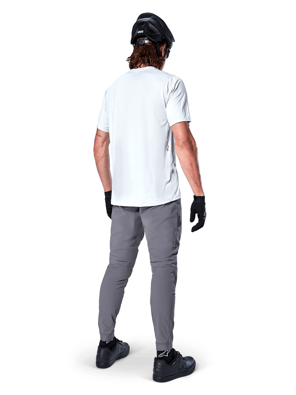 Alpinestars A-Dura byxor, mörkgrå MTB-byxor sedda bakifrån, bärs med en vit kortärmad t-shirt, svart MTB-hjälm, svarta handskar och skor, normal passform med elastiska muddar, slitstark utrustning för terrängcykling