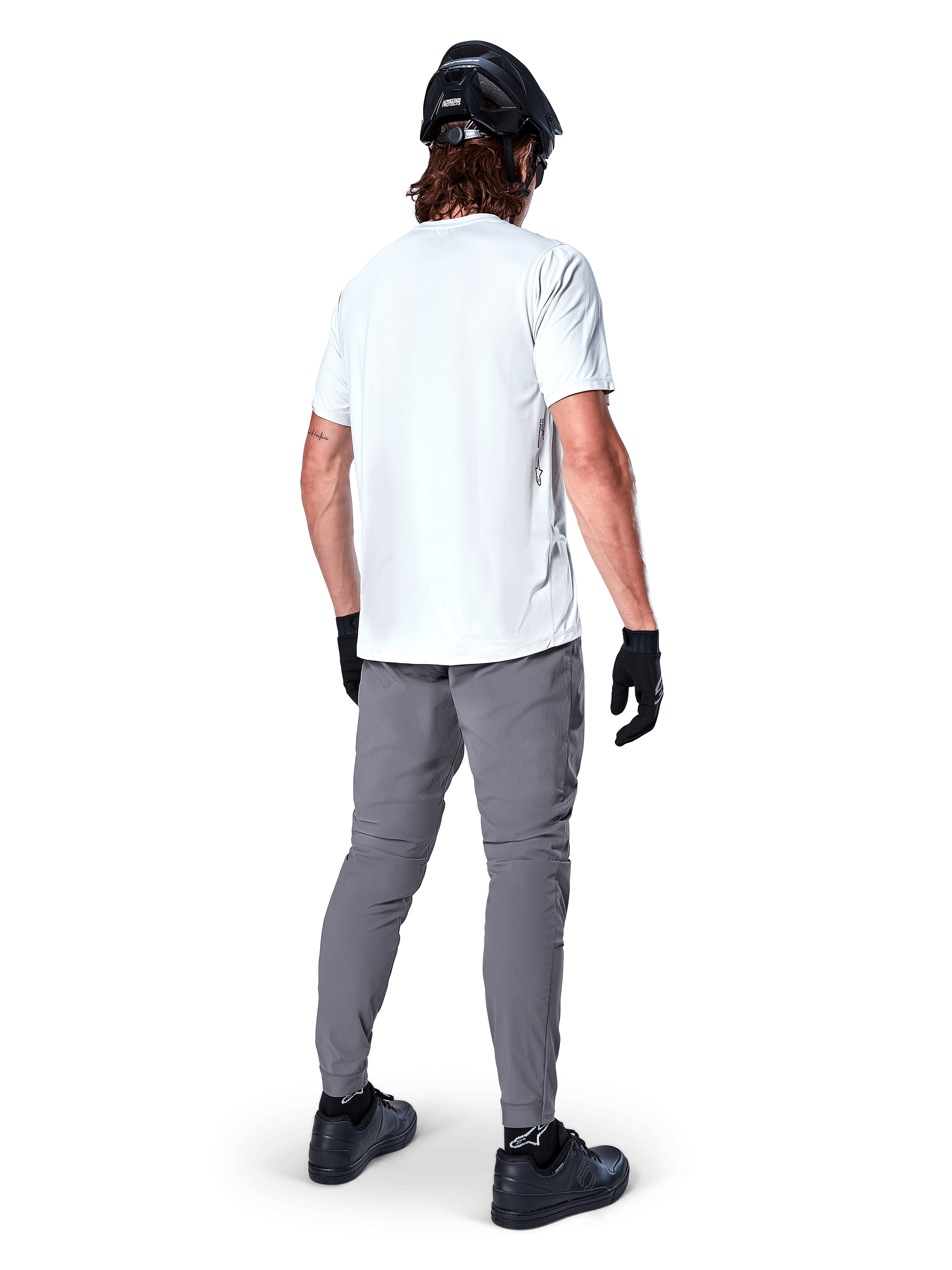 Alpinestars A-Dura byxor, mörkgrå MTB-byxor sedda bakifrån, bärs med en vit kortärmad t-shirt, svart MTB-hjälm, svarta handskar och skor, normal passform med elastiska muddar, slitstark utrustning för terrängcykling