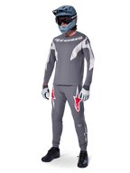 Alpinestars A-Supra Race Pants in Dark Gray, featuring bold red and white logos on the thighs, worn by a rider as part of a complete mountain bike racing kit with a matching gray jersey, blue-gray full-face helmet, and light gray gloves, high-performance gear for MTB racing