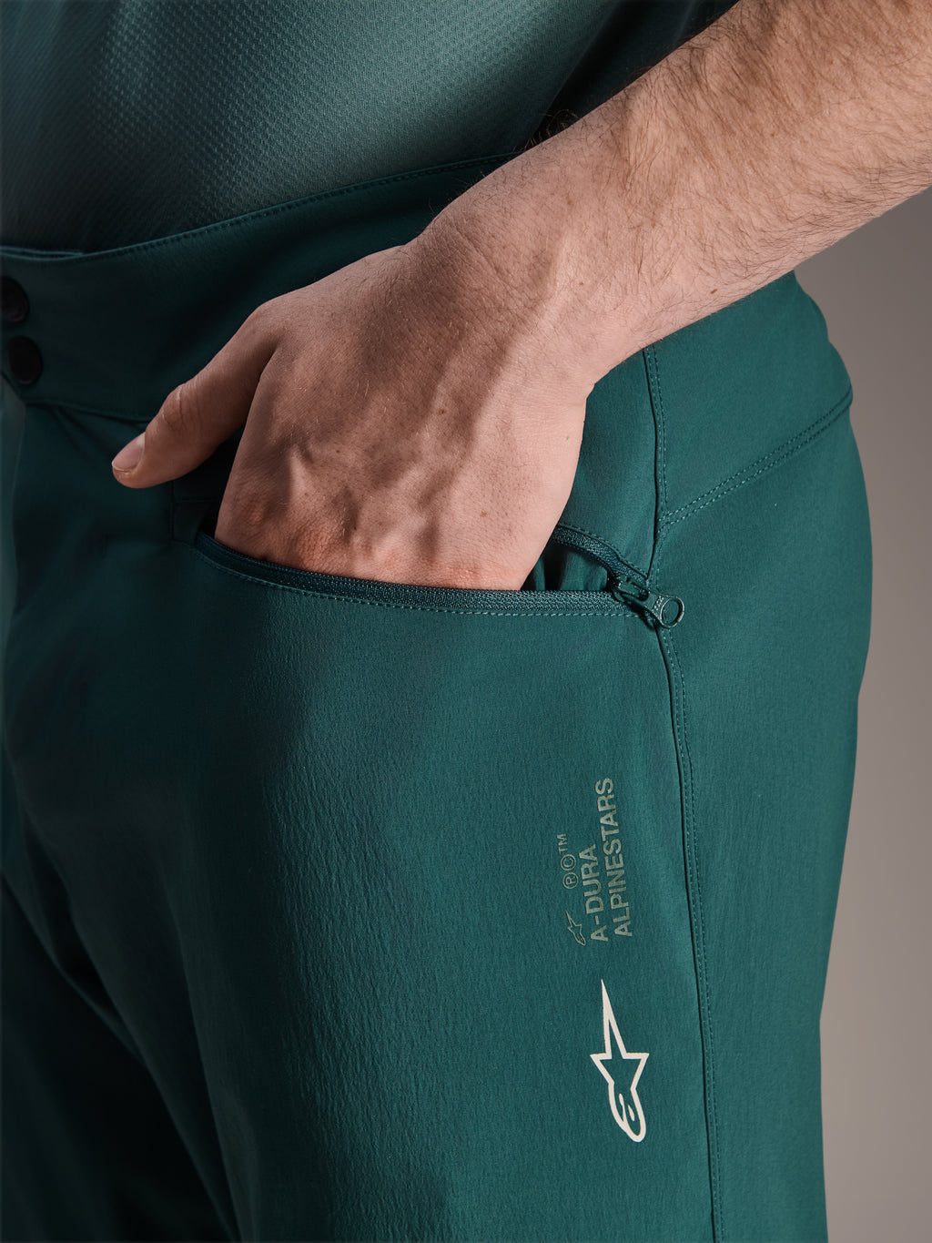 Alpinestars A-Dura Shorts i Dark Pine, närbild som visar en hand i den dragkedjeförsedda meshfodrade fickan, med vit Alpinestars stjärnlogotyp och A-Dura-branding, hållbar textilkonstruktion för mountainbike