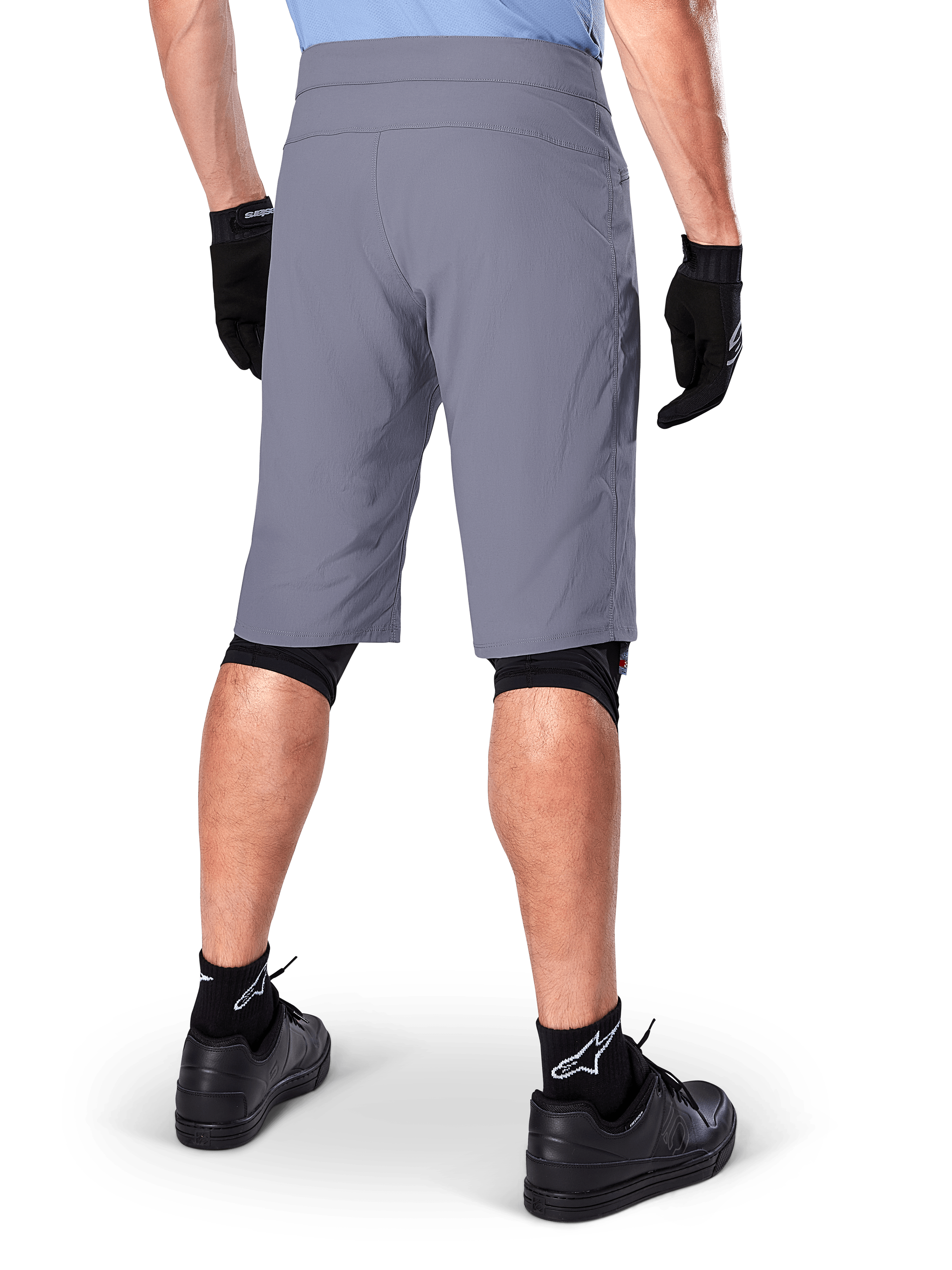 Alpinestars A-Dura Shorts, mörkgrå mountainbikeshorts, sedda bakifrån på en åkare, bärs med svarta handskar, svarta knäskyddsfoder och svarta strumpor med Alpinestars-logotyp, slitstark konstruktion för terrängcykling
