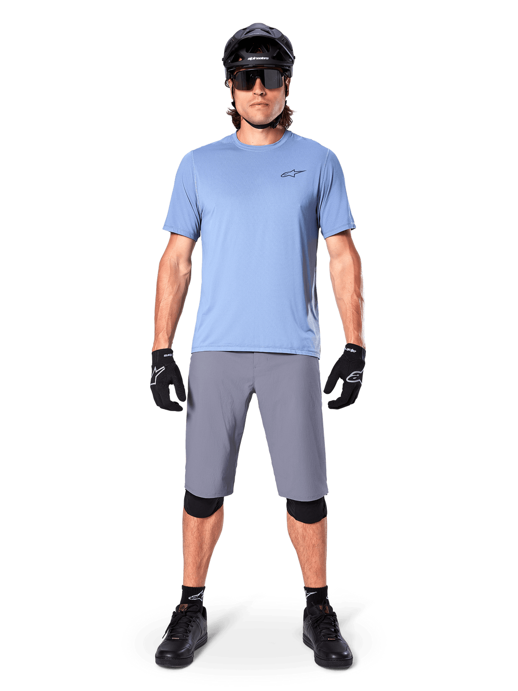 Manlig cyklist som bär Alpinestars A-Dura Shorts i mörkgrått, stylad med en ljusblå kortärmad t-shirt, svart hjälm, solglasögon och svarta knäskydd, komplett mountainbike-outfit för terrängprestanda.