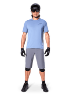 Manlig cyklist som bär Alpinestars A-Dura Shorts i mörkgrått, stylad med en ljusblå kortärmad t-shirt, svart hjälm, solglasögon och svarta knäskydd, komplett mountainbike-outfit för terrängprestanda.