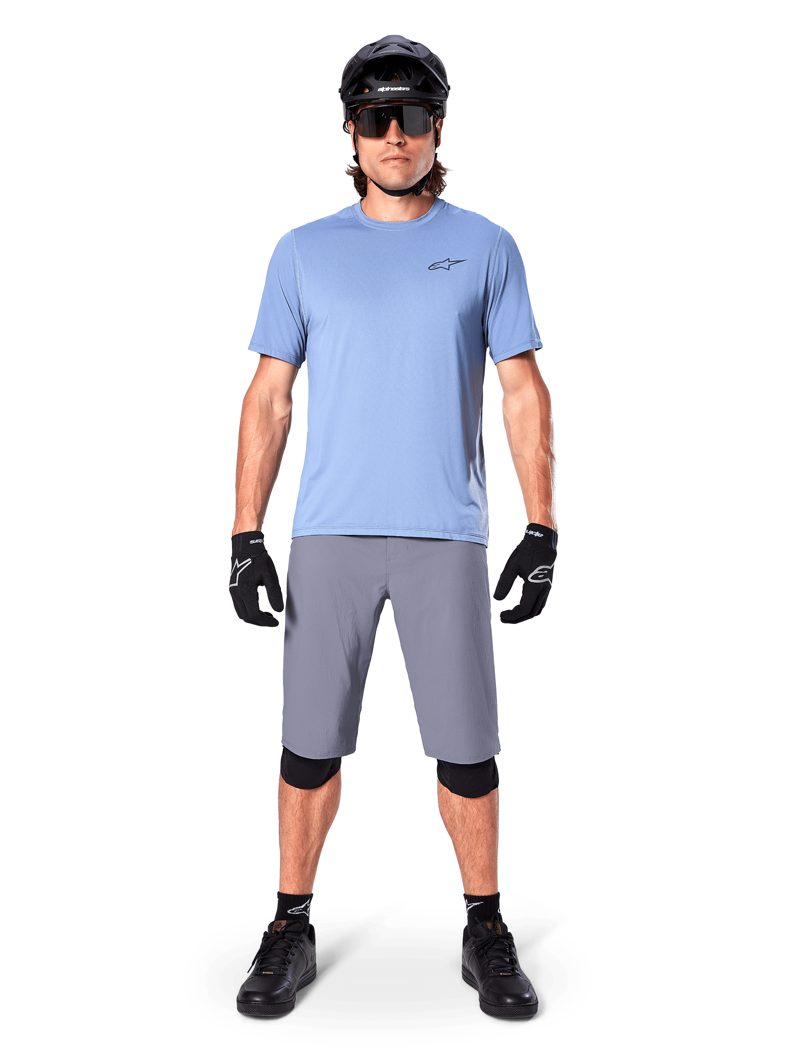 Manlig cyklist som bär Alpinestars A-Dura Shorts i mörkgrått, stylad med en ljusblå kortärmad t-shirt, svart hjälm, solglasögon och svarta knäskydd, komplett mountainbike-outfit för terrängprestanda.