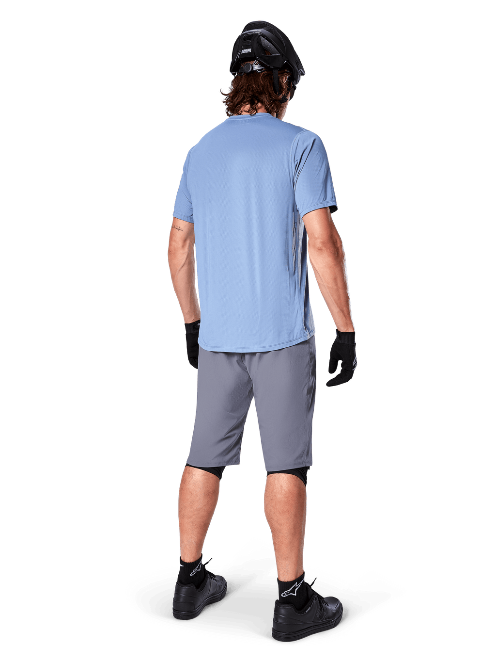 Alpinestars A-Dura Shorts, mörkgrå MTB-shorts, sedda bakifrån, bärs med en ljusblå kortärmad tröja och svart mountainbikehjälm, svarta handskar och strumpor med svarta skor, funktionell och slitstark utrustning för terrängcykling