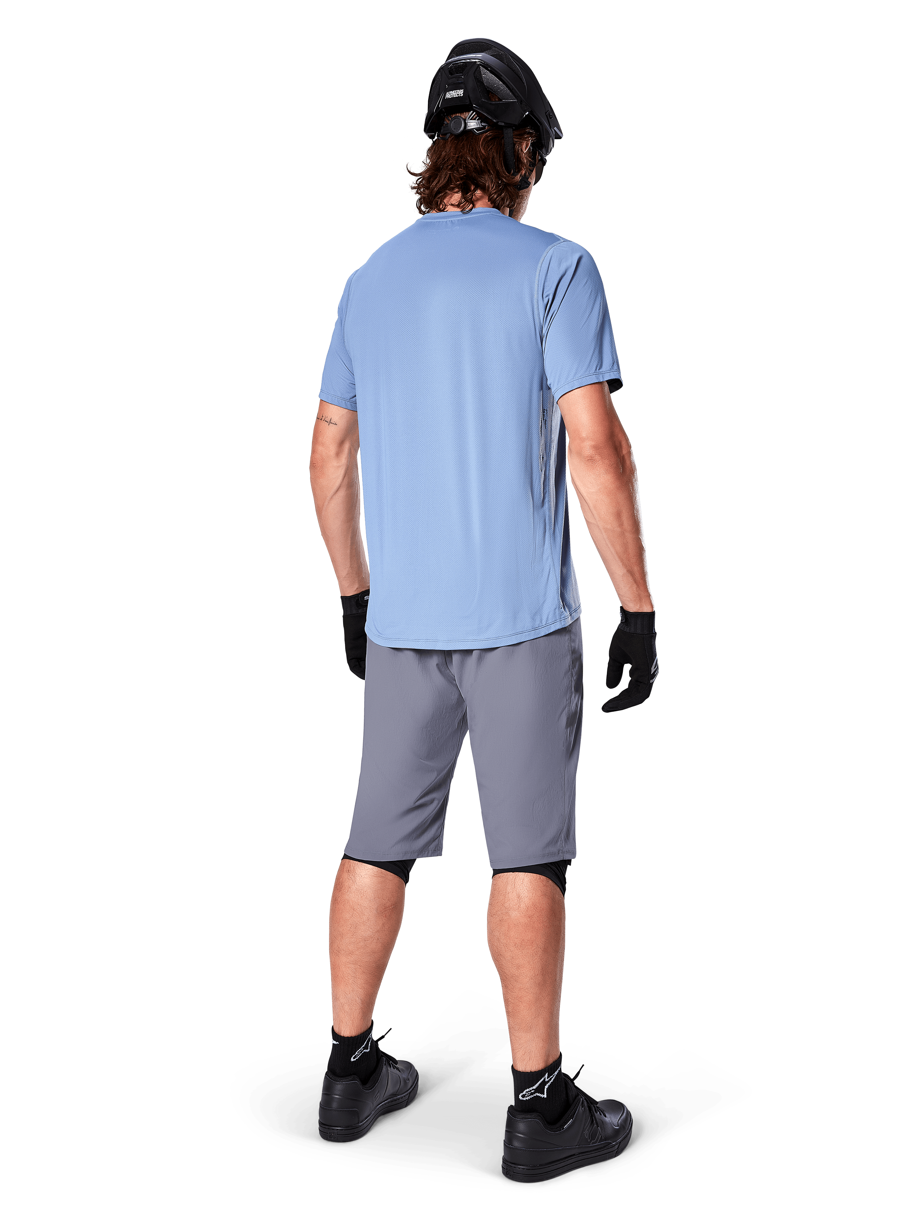 Alpinestars A-Dura Shorts, mörkgrå MTB-shorts, sedda bakifrån, bärs med en ljusblå kortärmad tröja och svart mountainbikehjälm, svarta handskar och strumpor med svarta skor, funktionell och slitstark utrustning för terrängcykling