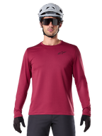Manlig cyklist som bär Alpinestars A-Dura Thermal Jersey, långärmad MTB-tröja, vinröd, matchad med en grå hjälm och svarta solglasögon, har en liten svart stjärnlogotyp på bröstet, isolerat fleecetyg för mountainbike-turer i kallt väder