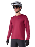 Alpinestars A-Dura Thermal Jersey, långärmad mountainbiketröja, vinröd, med isolerande fleecefoder och en liten svart logotyp på bröstet, bärs med en grå hjälm, svarta solglasögon och svarta handskar, designad för cykling i kallt väder