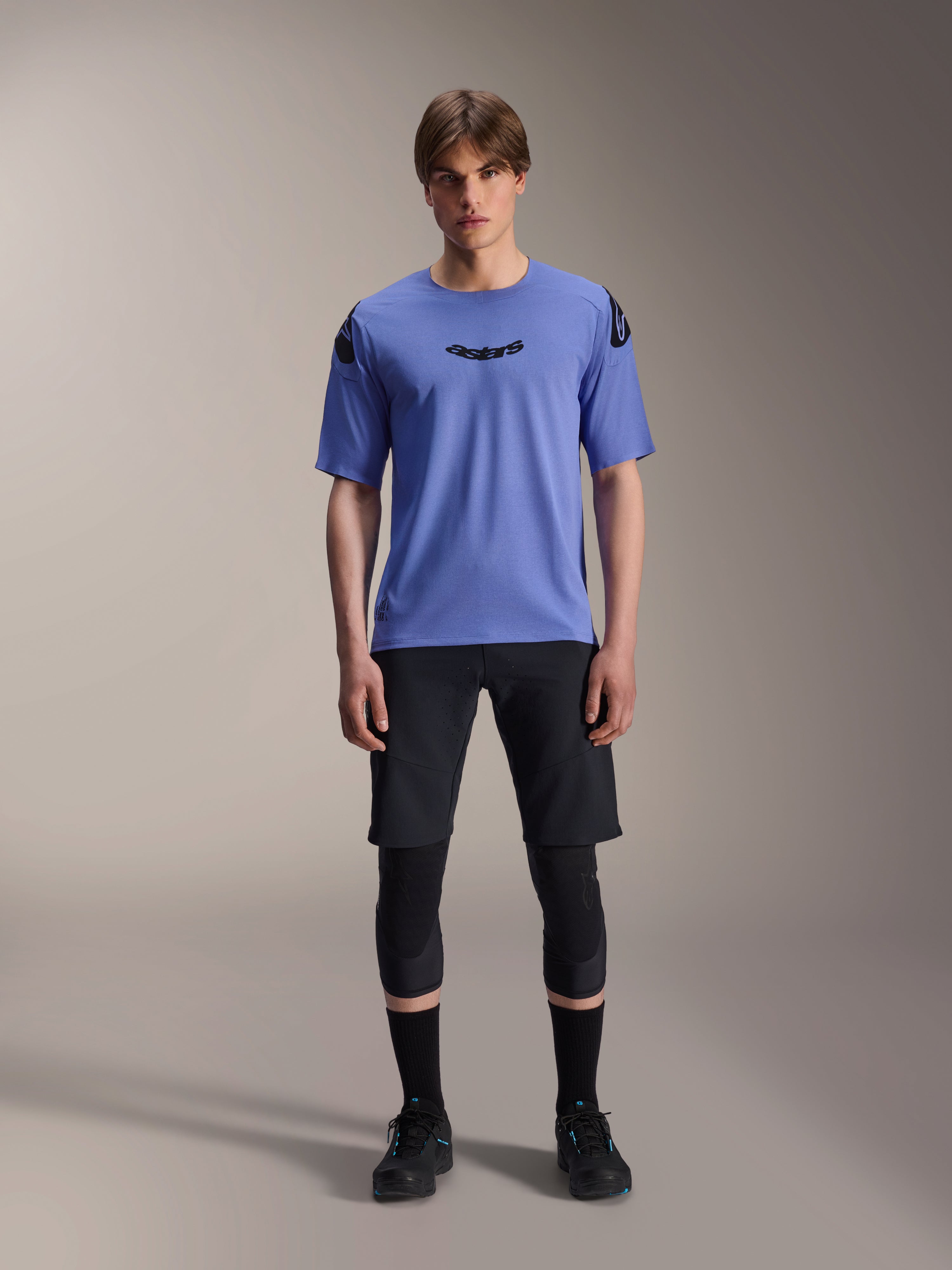 Alpinestars A-Aria Elite Shorts, MTB Mountain Biking Shorts, Black Titanium, worn with blue short-sleeve jersey, knee guards, and black biking shoes, full-body studio shot, performance mountain bike apparel