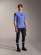 Manlig modell som bär Alpinestars A-Aria Elite Shorts i Black Titanium och A-Aria Polartec Elite SS Jersey i ljuslila, matchat med svarta knäskydd för mountainbike och skor, professionell cykelutrustning för off-road-prestanda.