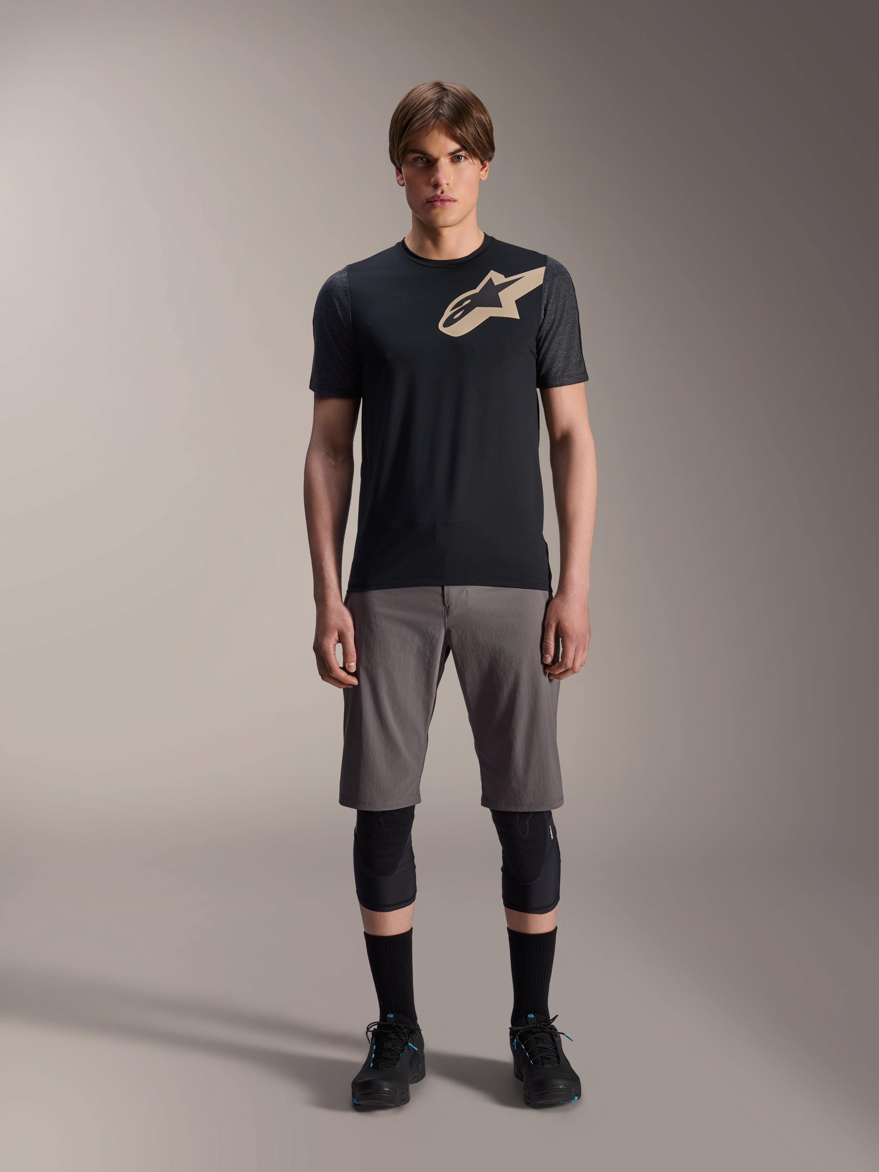 Alpinestars A-Dura Lipan kortärmad tröja, svart med gråmelerade ärmar och beige Astars-logotyp på bröstet, bärs av en manlig modell med grå mountainbikeshorts och svarta knäskydd, funktionell MTB-utrustning.