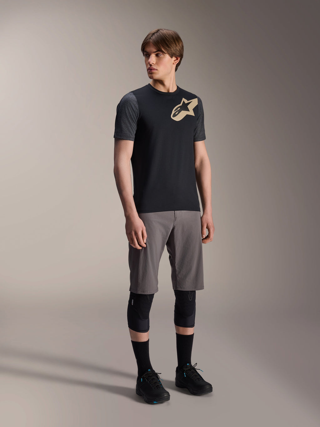 Manlig modell i Alpinestars A-Dura Lipan kortärmad tröja i svart, med mörkgrå ärmar och en beige Astars-logotyp på bröstet, kombinerat med grå mountainbike-shorts, svarta knäskydd och svarta cykelskor, koordinerad mountainbike-utrustning