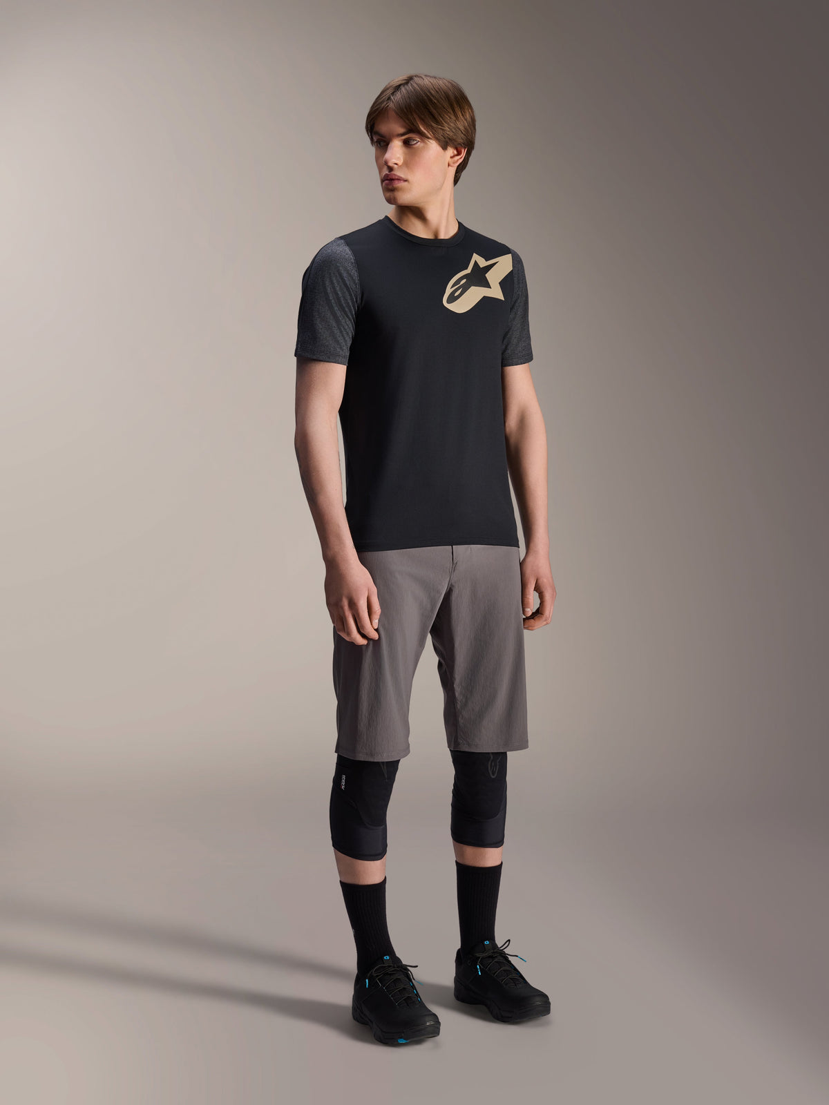 Manlig modell i Alpinestars A-Dura Lipan kortärmad tröja i svart, med mörkgrå ärmar och en beige Astars-logotyp på bröstet, kombinerat med grå mountainbike-shorts, svarta knäskydd och svarta cykelskor, koordinerad mountainbike-utrustning