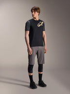 Manlig modell i Alpinestars A-Dura Lipan kortärmad tröja i svart, med mörkgrå ärmar och en beige Astars-logotyp på bröstet, kombinerat med grå mountainbike-shorts, svarta knäskydd och svarta cykelskor, koordinerad mountainbike-utrustning