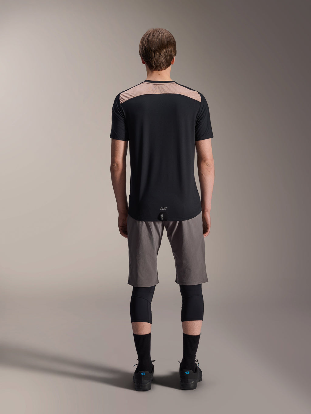 Alpinestars A-Dura Lipan tröja - kortärmad, svart med beige övre ryggpanel, vy bakifrån som visar förlängd bakdel, bärs med gråa MTB-shorts och svarta knäskydd, prestandakläder för mountainbike