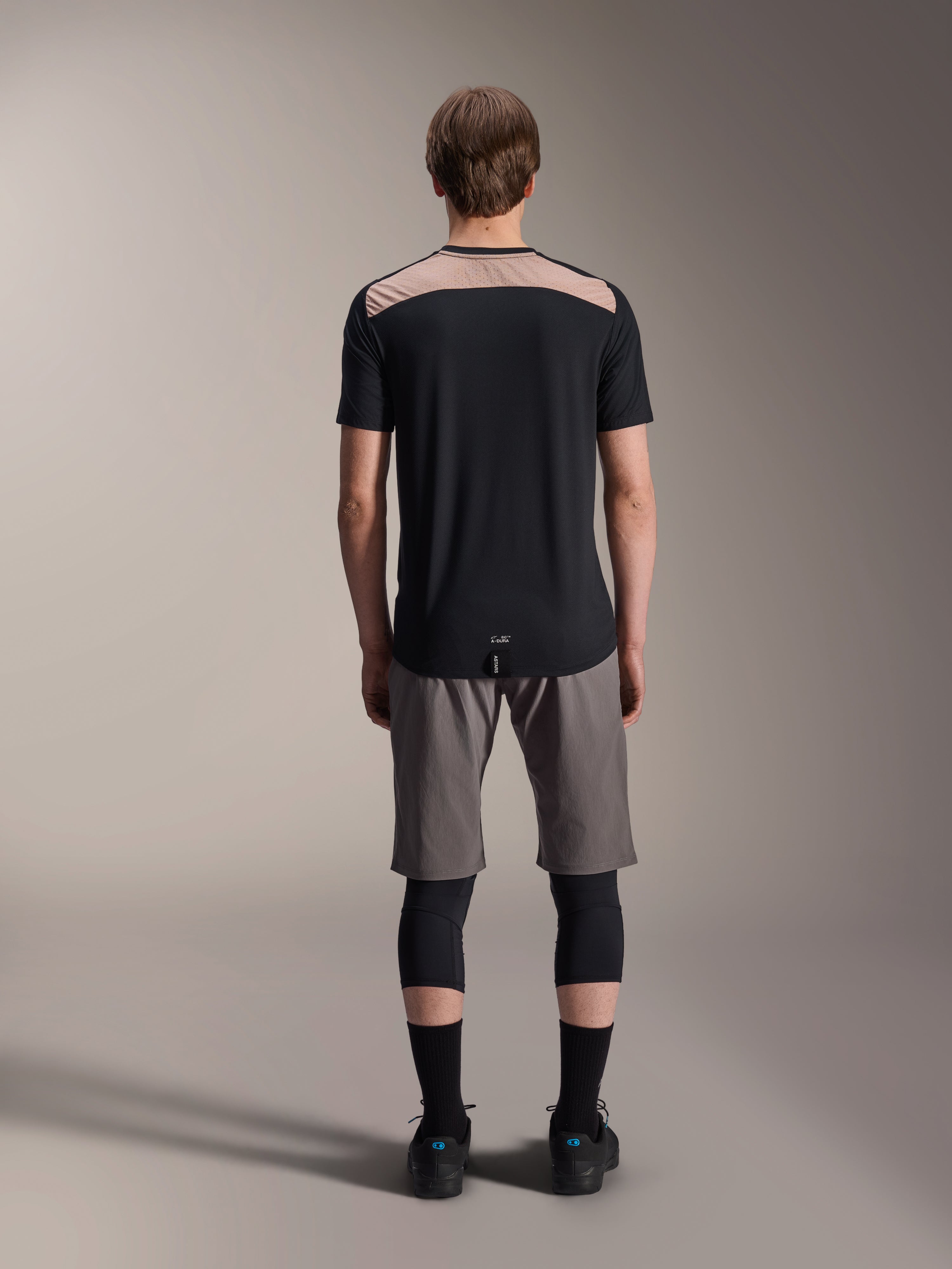 Alpinestars A-Dura Lipan tröja - kortärmad, svart med beige övre ryggpanel, vy bakifrån som visar förlängd bakdel, bärs med gråa MTB-shorts och svarta knäskydd, prestandakläder för mountainbike