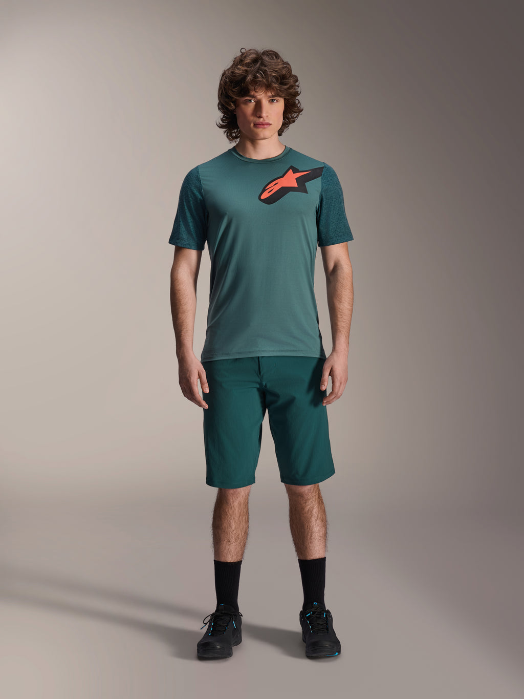 Alpinestars A-Dura Shorts i Dark Pine, MTB-shorts för herr, visas på en modell med en matchande mörkblågrön kortärmad tröja med röd och svart stjärnlogotyp, svarta strumpor och cykelskor, avslappnad sportig mountainbike-klädsel