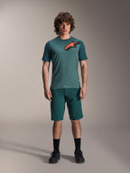 Alpinestars A-Dura Shorts i Dark Pine, MTB-shorts för herr, visas på en modell med en matchande mörkblågrön kortärmad tröja med röd och svart stjärnlogotyp, svarta strumpor och cykelskor, avslappnad sportig mountainbike-klädsel