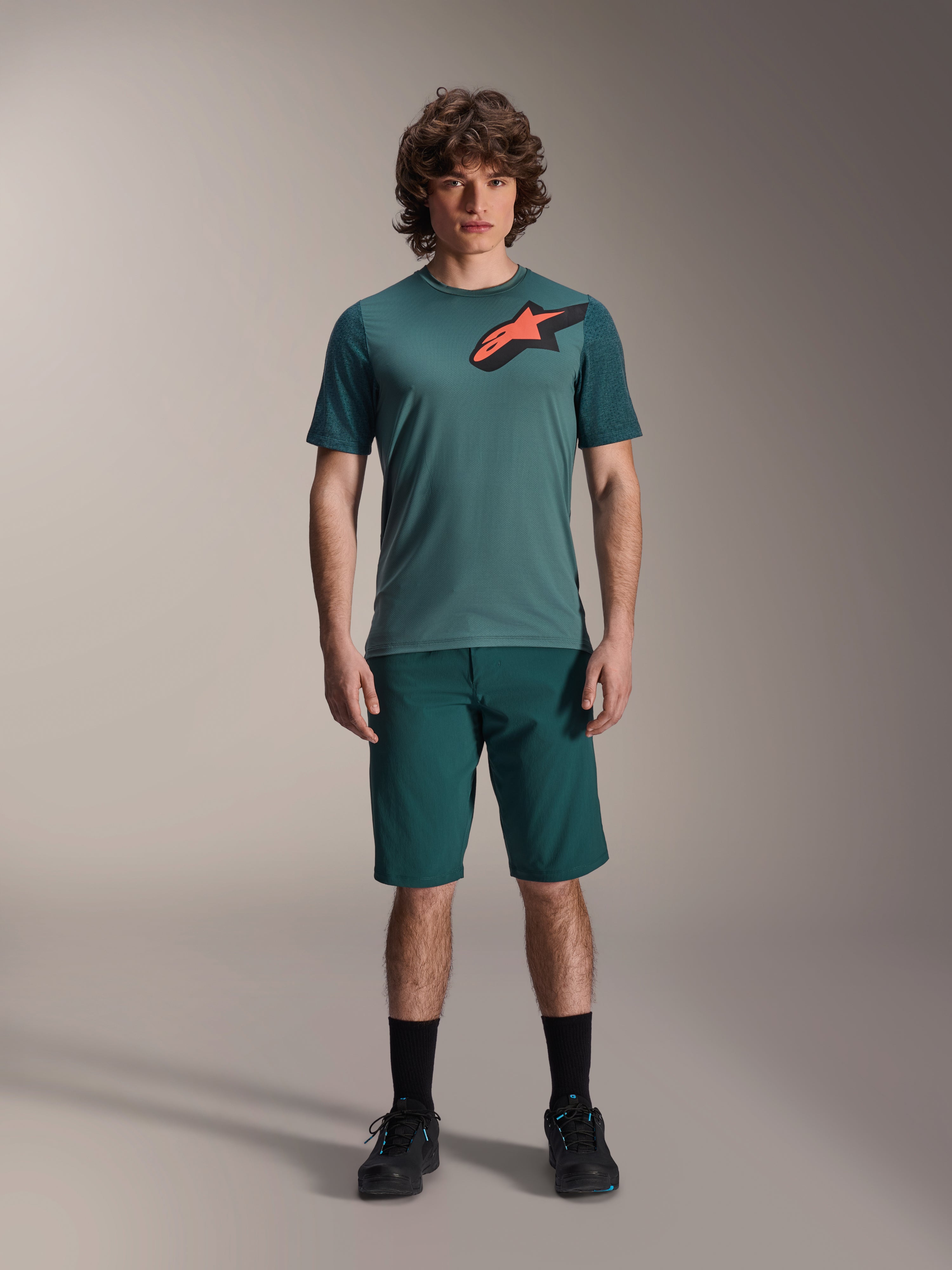 Alpinestars A-Dura Shorts i Dark Pine, MTB-shorts för herr, visas på en modell med en matchande mörkblågrön kortärmad tröja med röd och svart stjärnlogotyp, svarta strumpor och cykelskor, avslappnad sportig mountainbike-klädsel