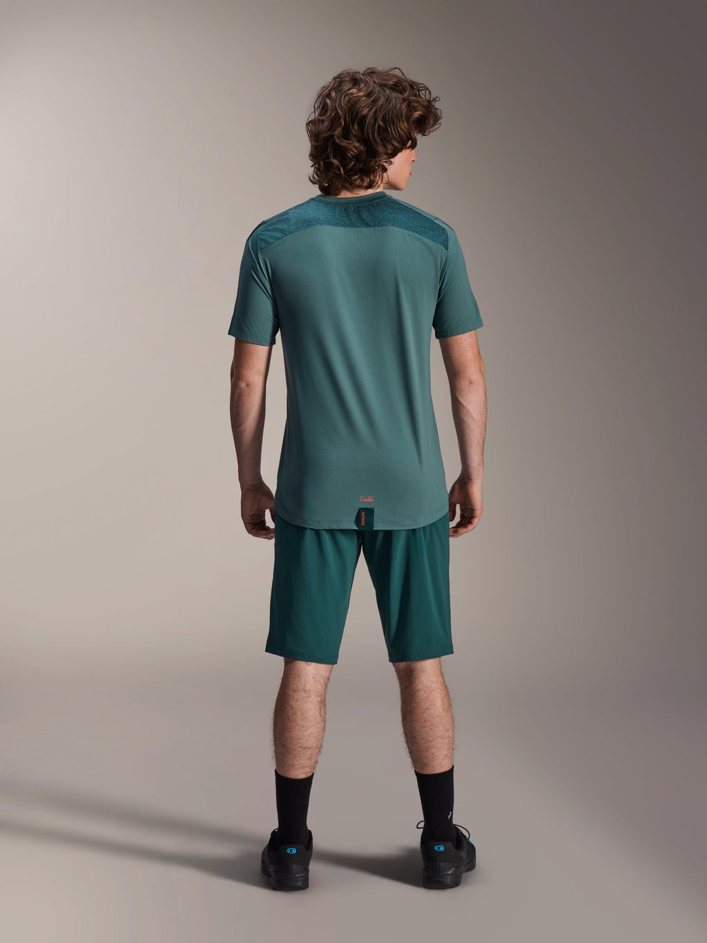 Alpinestars A-Dura Lipan kortärmad tröja i Silver Pine, vy bakifrån som visar de texturerade jacquard-axelpanelerna och det förlängda bakstycket, buren med matchande mörkgröna mountainbikeshorts och svarta skor.