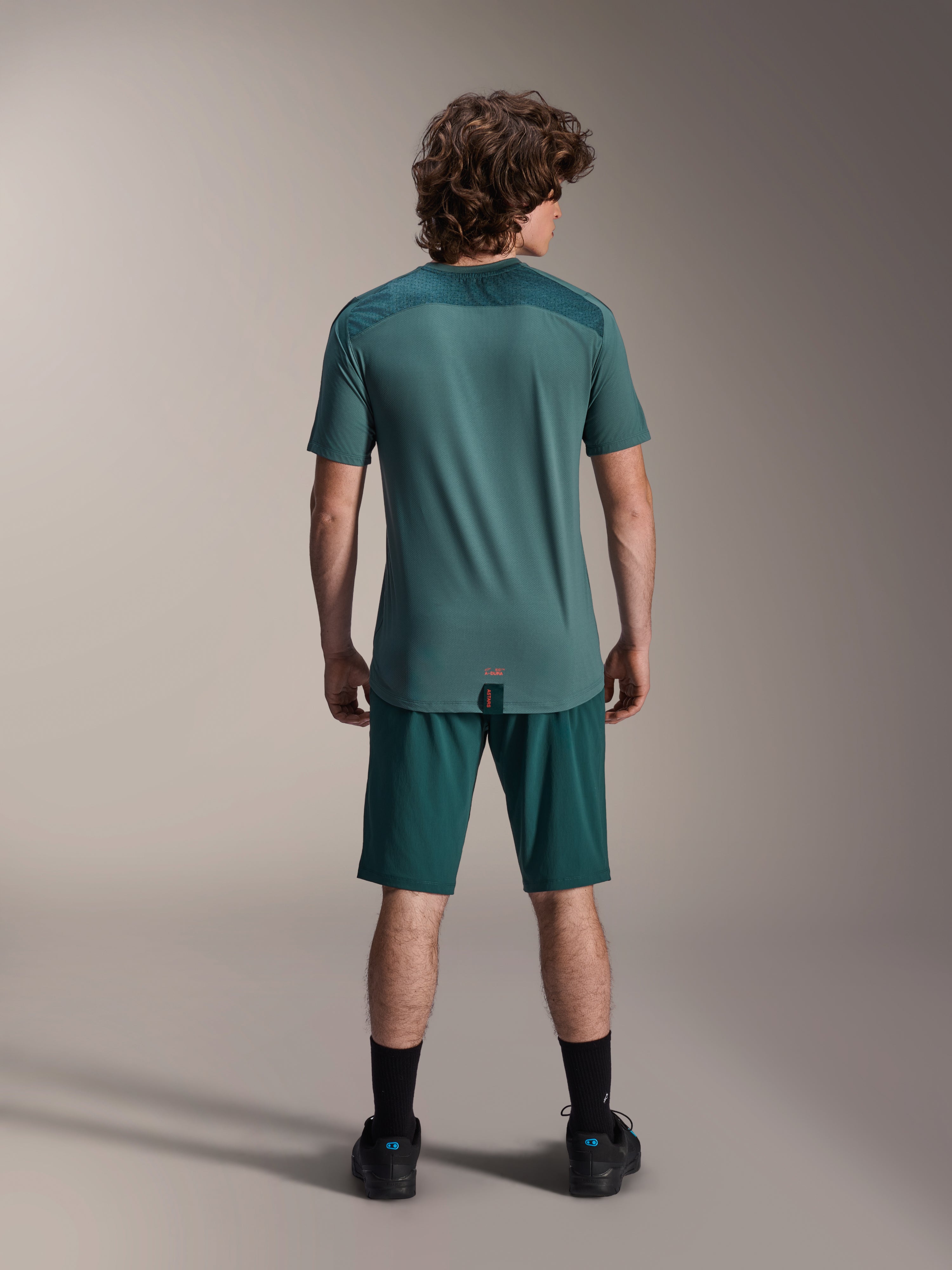 Alpinestars A-Dura Lipan kortärmad tröja i Silver Pine, vy bakifrån som visar de texturerade jacquard-axelpanelerna och det förlängda bakstycket, buren med matchande mörkgröna mountainbikeshorts och svarta skor.