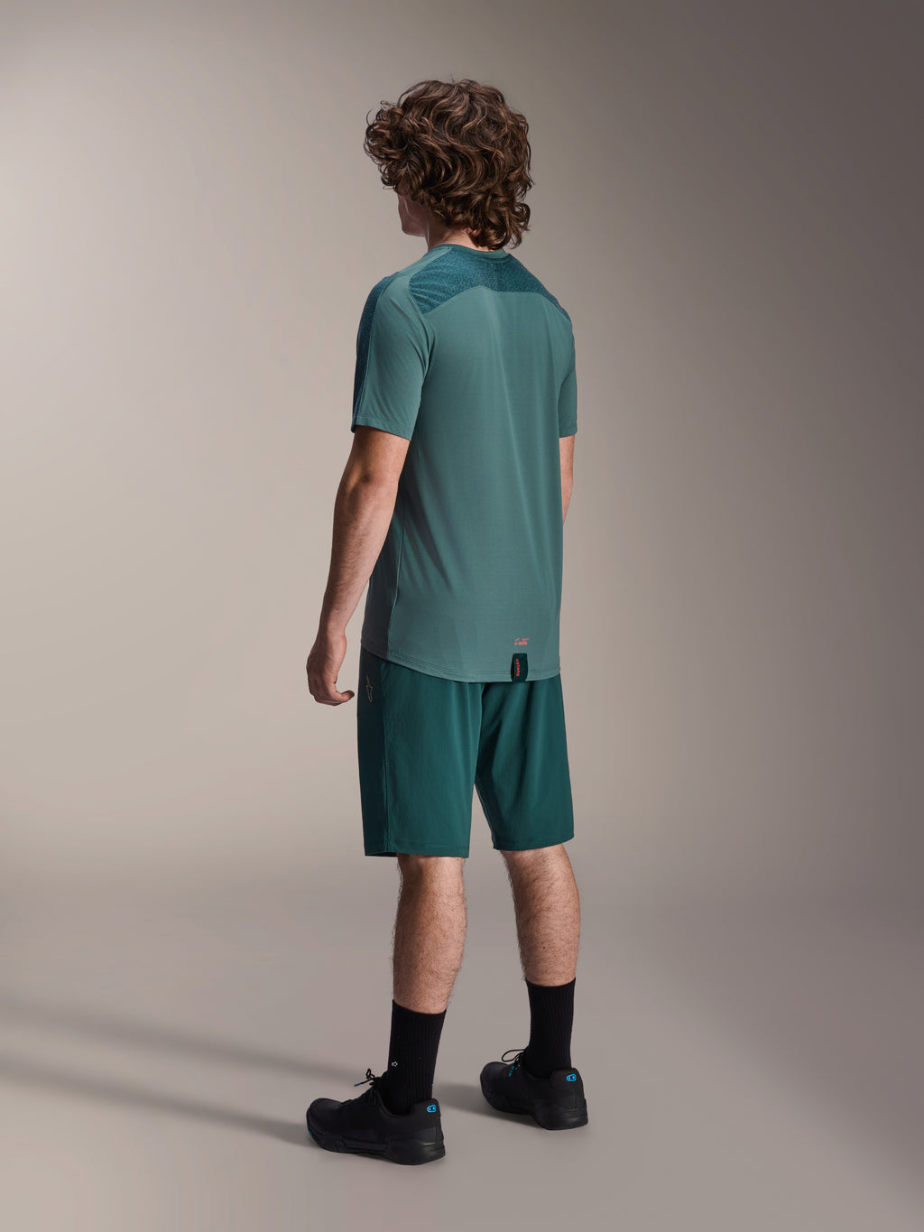 Modell bär Alpinestars A-Dura Shorts i Dark Pine och matchande grön kortärmad Lipan-tröja, sedd bakifrån, kombinerat med svarta strumpor och svarta mountainbikeskor, professionell MTB-utrustning.