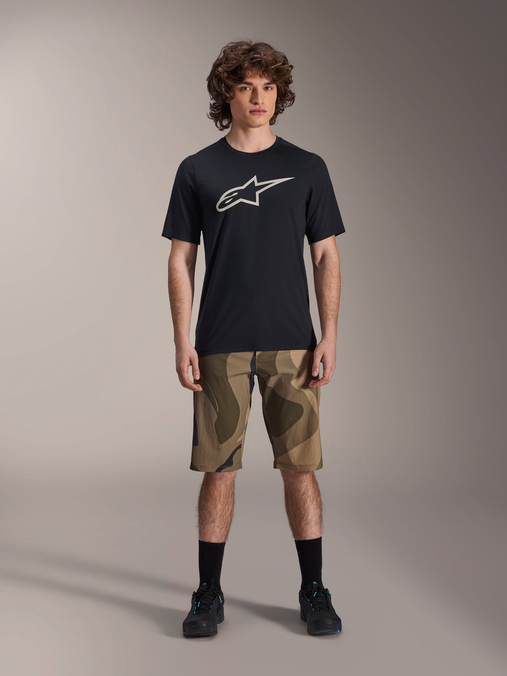 Alpinestars A-Dura Astar kortärmad tröja, svart med stor vit Astars-logotyp på bröstet, bärs av en modell med kamouflagemönstrade MTB-shorts och svarta cykelskor, avslappnade mountainbike-kläder.