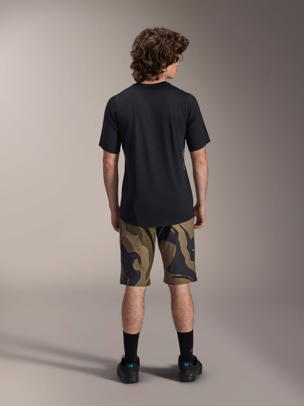 Alpinestars A-Dura Astar kortärmad tröja, svart, vy bakifrån på en modell kombinerad med kamouflagemönstrade MTB-shorts och svarta skor, avslappnade tekniska mountainbike-kläder