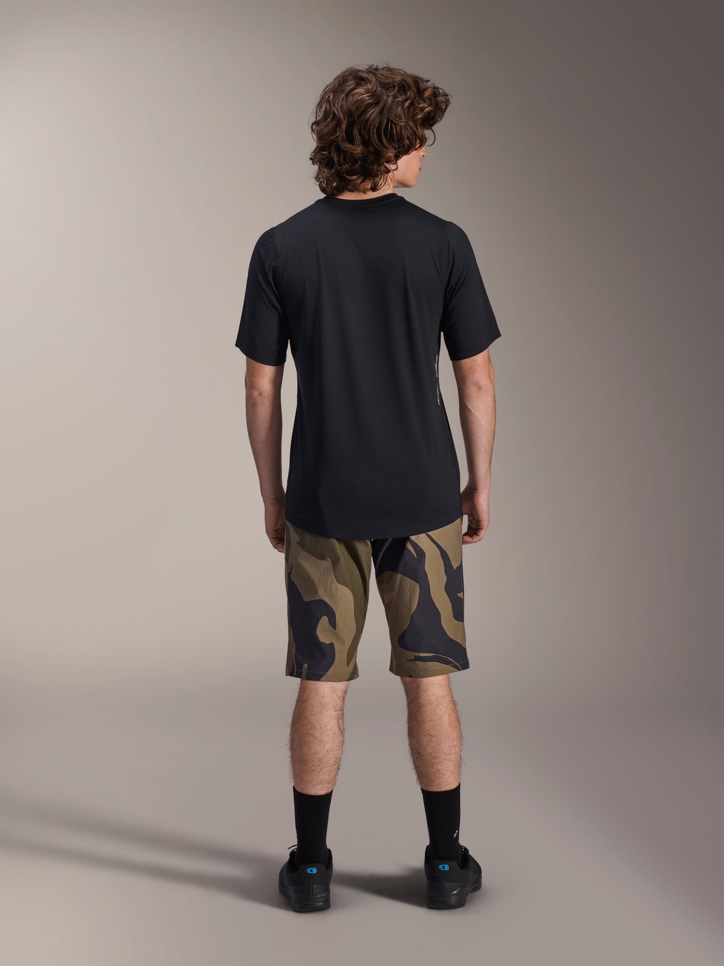 Alpinestars A-Dura Astar kortärmad tröja, svart, vy bakifrån på en modell kombinerad med kamouflagemönstrade MTB-shorts och svarta skor, avslappnade tekniska mountainbike-kläder