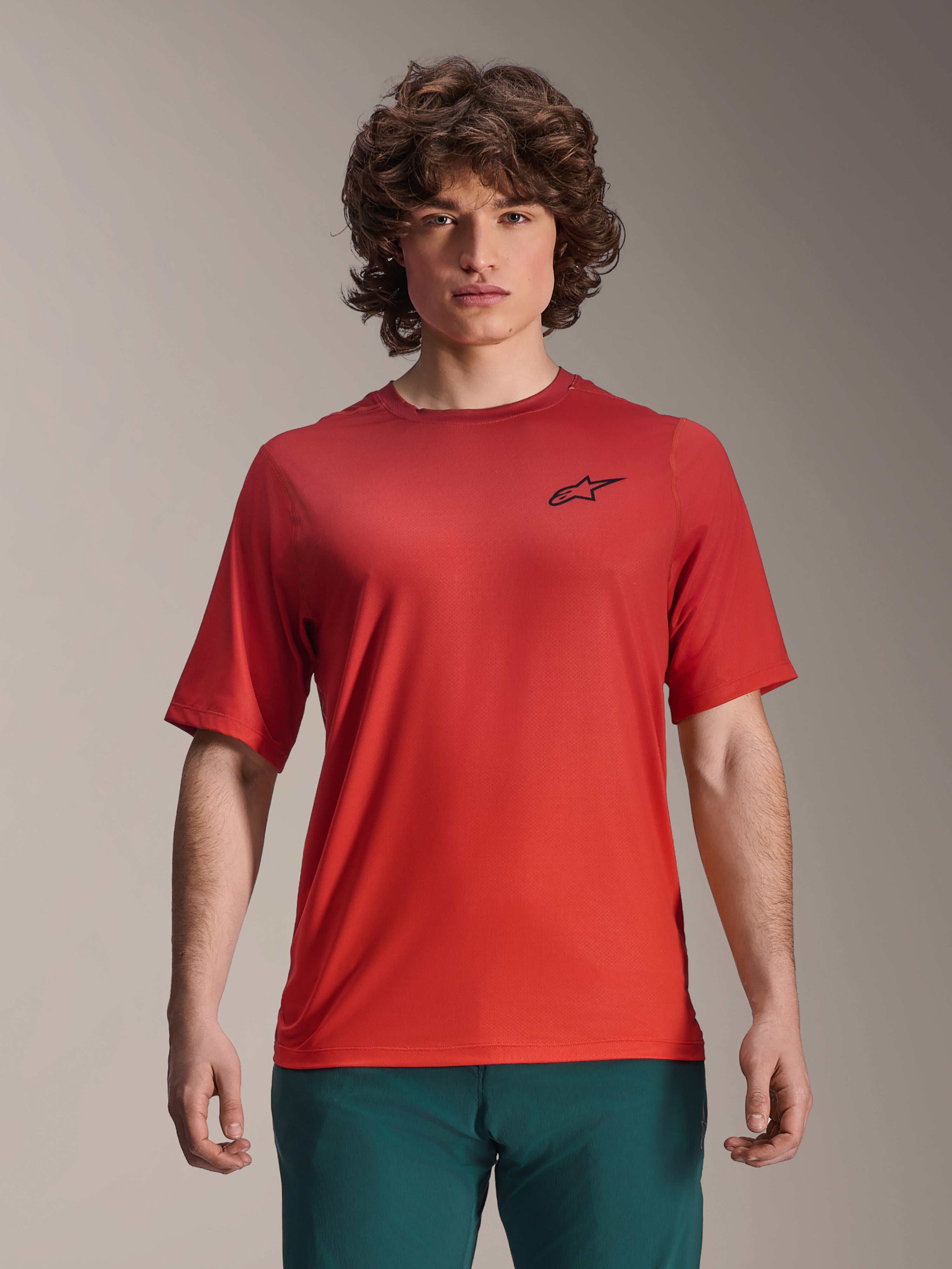 Alpinestars 2025 A-Dura Rise Jersey, kortärmad MTB-tröja, Spicy Orange, bärs av en man, med en svart Astars-logotyp på bröstet, kombinerad med mörkgröna byxor, avslappnad funktionsklädsel för mountainbike