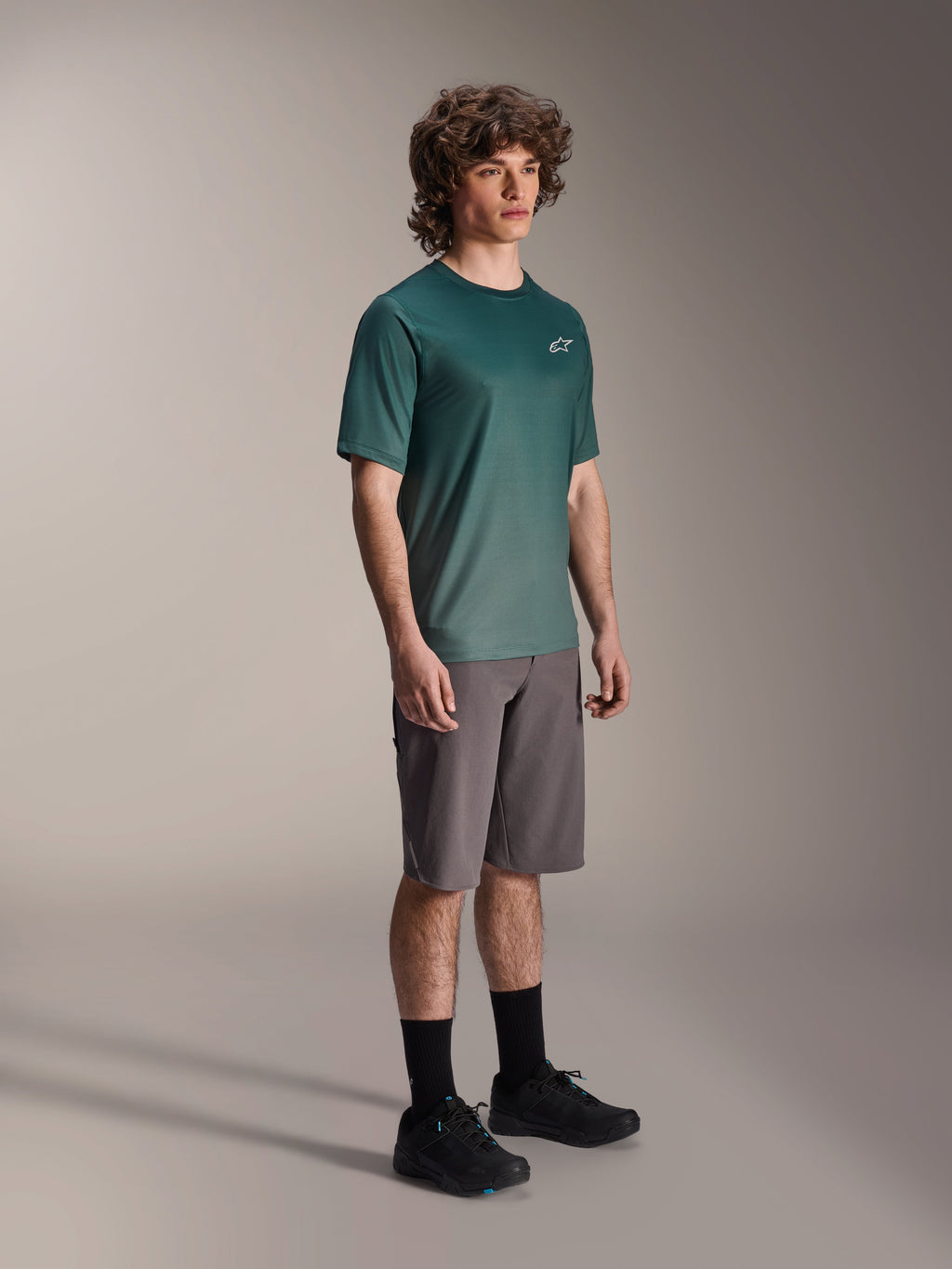 Alpinestars A-Dura Rise Jersey - Short Sleeve i Silver Pine, grön mountainbiketröja bärs av en modell med gråa shorts och svarta sneakers, avslappnad MTB-utrustning för stigcykling med en liten vit logotyp på bröstet