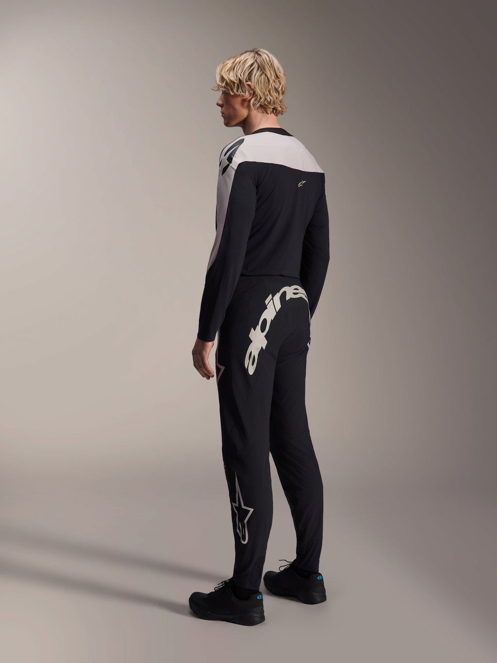 Alpinestars A-Supra Melt långärmad tröja, svart med vita axelpaneler, bärs av en modell bakifrån och från sidan, kombinerad med svarta mountainbikebyxor med ett stort vitt ordmärke, högpresterande MTB-racingkit