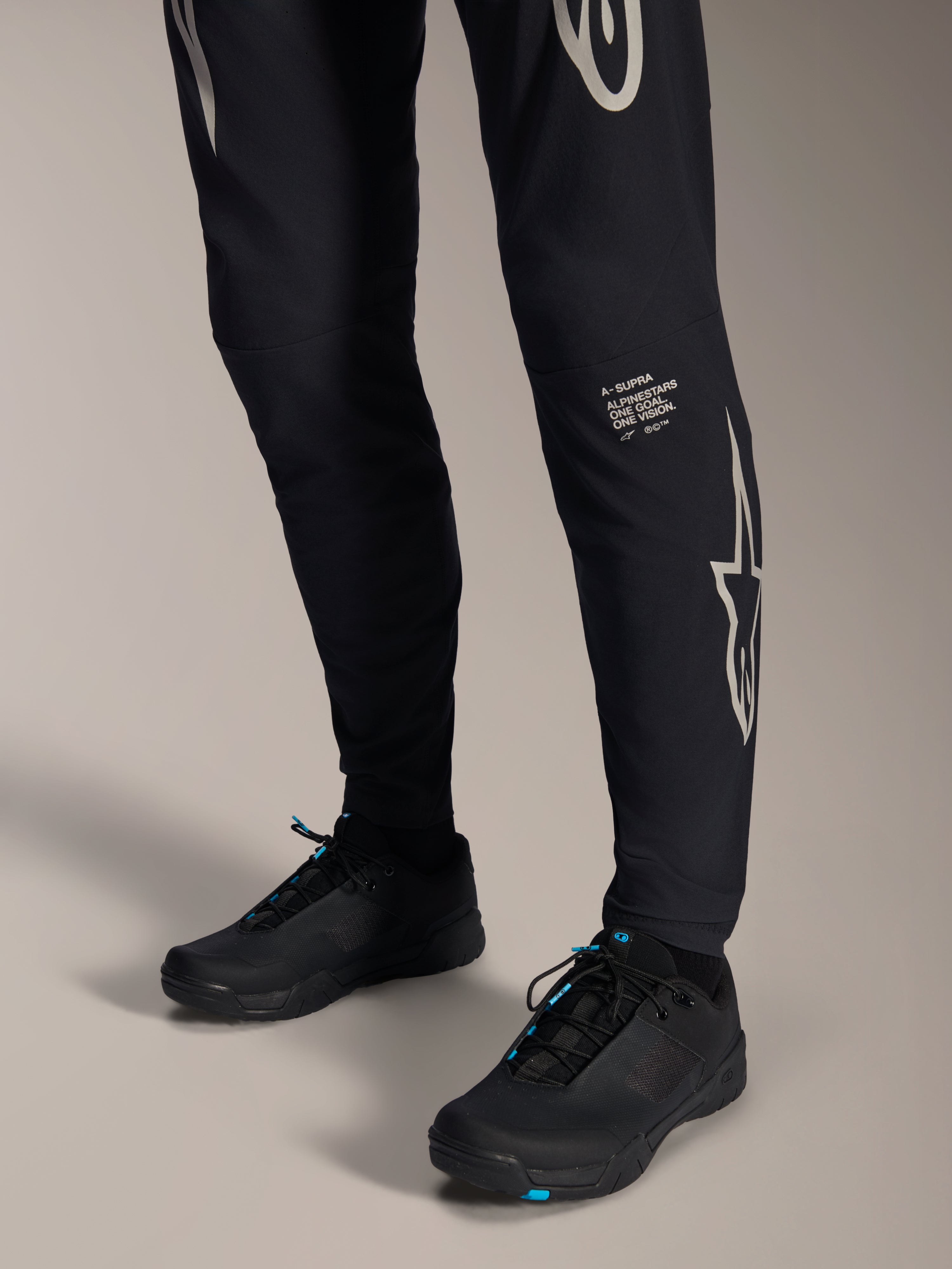 Alpinestars A-Supra Melt-byxor, svarta mountainbikebyxor, med vitt Alpinestars-ordmärke och logotyp på benet, bärs med svarta cykelskor med blå detaljer, högpresterande terrängutrustning