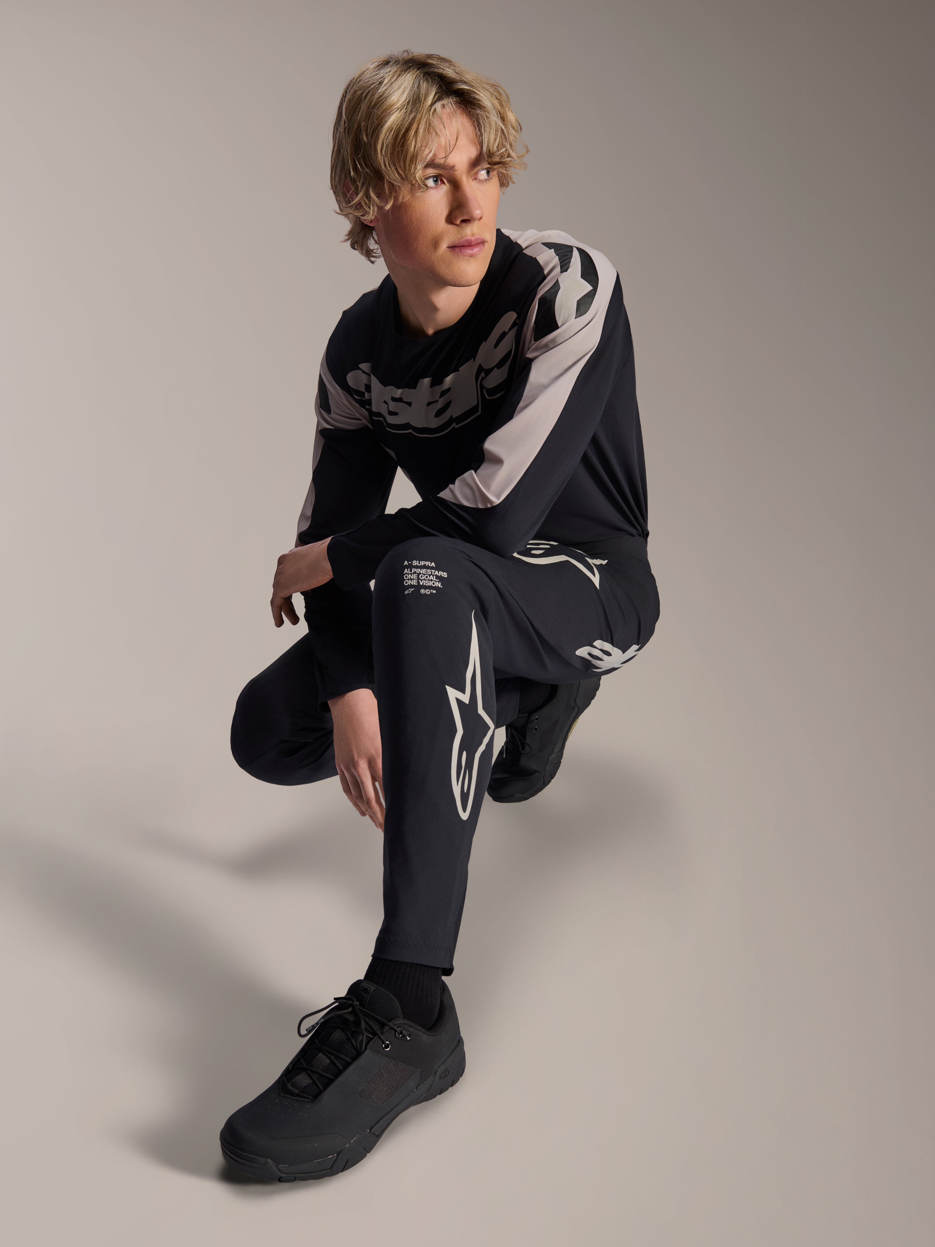 Alpinestars A-Supra Melt-byxor och långärmad tröja, svarta och grå, med framträdande vit branding och stjärnlogotyper, buren av en manlig modell i en hukande pose, matchande MTB-racingutrustning