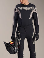 Alpinestars A-Supra Melt långärmad tröja, svart med grå grafiska paneler och framträdande 'astars'-logotyp på bröstet, bärs med svarta mountainbikebyxor och handskar medan personen håller en svart fullfacehjälm, racingutrustning för MTB