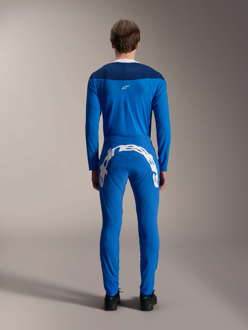 Manlig modell sedd bakifrån iförd A-Supra Melt Jersey - Long Sleeve i blått, mörkblå axelpanel med en liten vit stjärnlogotyp, matchande blå MTB-byxor med stor vit Alpinestars-logotyp över baken, koordinerad funktionell cykeloutfit för mountainbike.