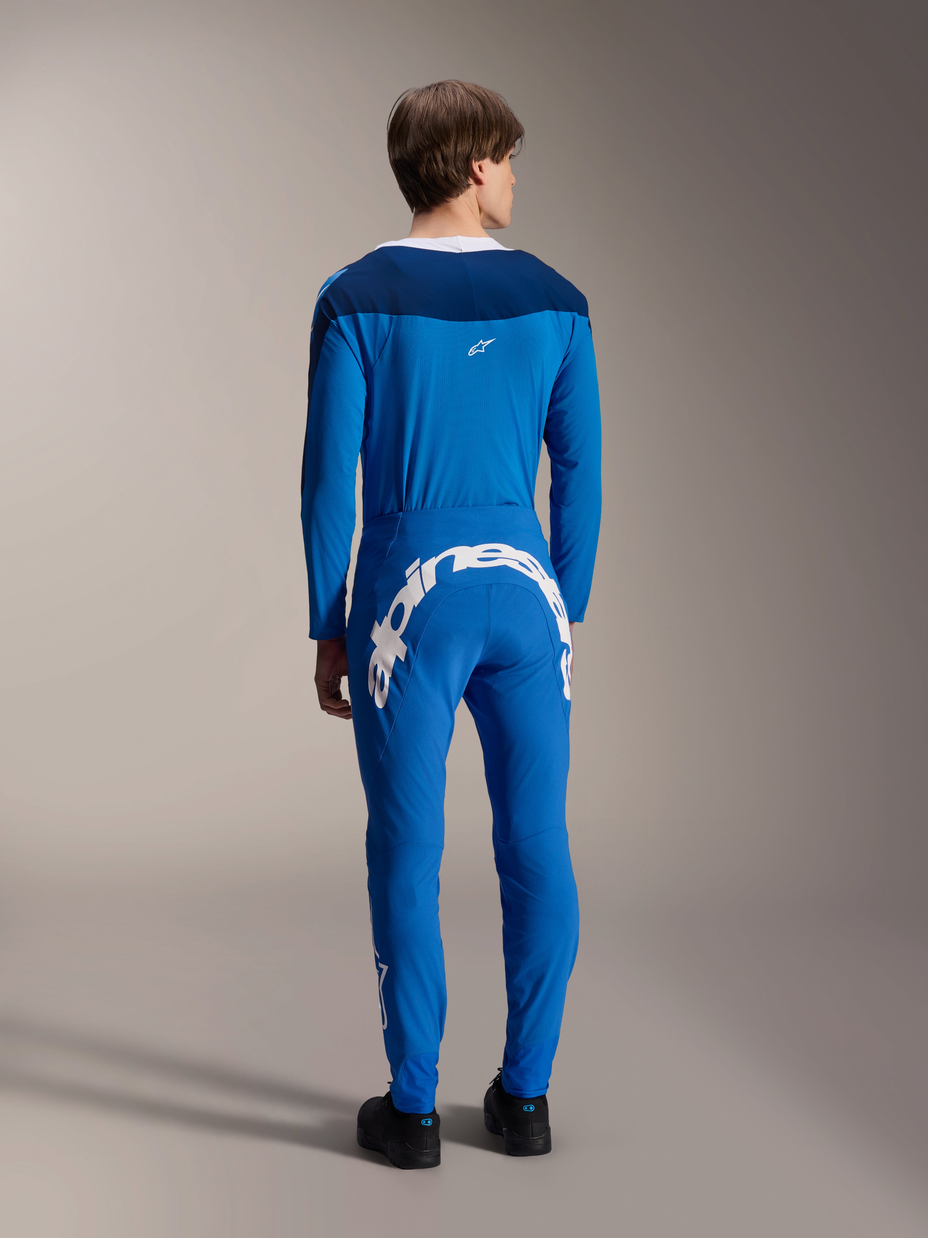 Manlig modell sedd bakifrån i Alpinestars A-Supra Melt långärmad tröja i blått, matchad med matchande blå funktionsbyxor med ett stort vitt Alpinestars-ordmärke över baksidan, komplett MTB-raceställ.
