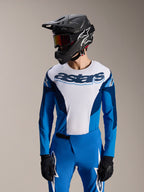 Modell iklädd Alpinestars A-Supra Melt långärmad tröja i blått och vitt med 'astars'-ordmärke på bröstet, stylad med matchande blå MTB-byxor, svart fullface-hjälm och goggles, komplett outfit för mountainbike-racing.
