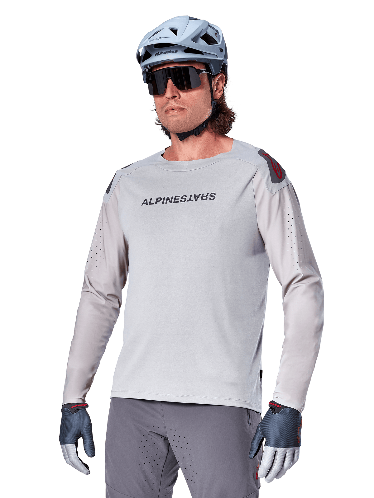 Alpinestars A-Aria Polartec Switch långärmad cykeltröja, ljusgrå med svart logotyp på bröstet och röd detalj på axeln, laserperforerade ärmar för luftflöde, bärs med en ljusblå hjälm, solglasögon och gråa byxor, funktionella mountainbikekläder för stigcykling