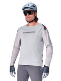 Alpinestars A-Aria Polartec Switch långärmad cykeltröja, ljusgrå med svart logotyp på bröstet och röd detalj på axeln, laserperforerade ärmar för luftflöde, bärs med en ljusblå hjälm, solglasögon och gråa byxor, funktionella mountainbikekläder för stigcykling