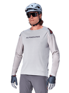 Alpinestars A-Aria Polartec Switch långärmad cykeltröja, ljusgrå med svart logotyp på bröstet och röd detalj på axeln, laserperforerade ärmar för luftflöde, bärs med en ljusblå hjälm, solglasögon och gråa byxor, funktionella mountainbikekläder för stigcykling