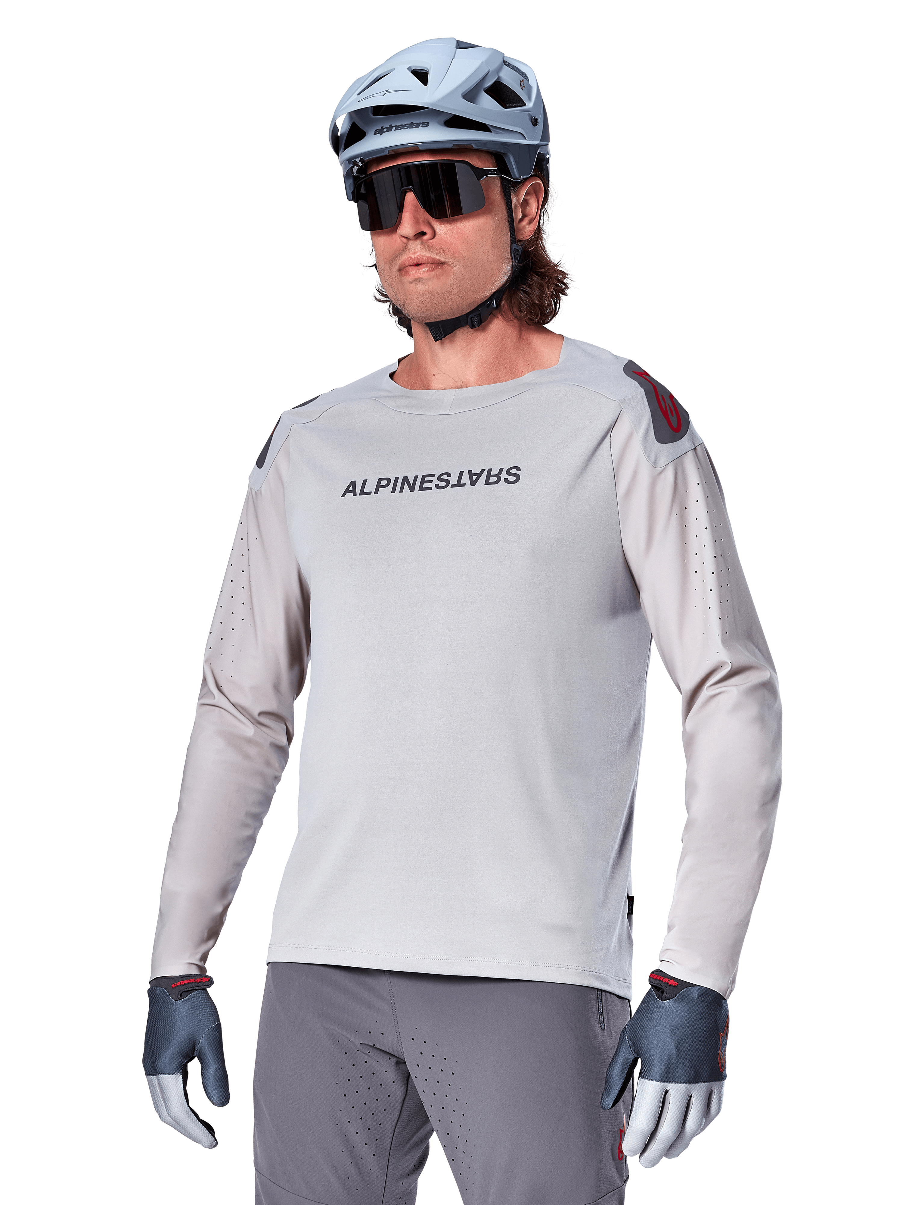 Alpinestars A-Aria Polartec Switch långärmad cykeltröja, ljusgrå med svart logotyp på bröstet och röd detalj på axeln, laserperforerade ärmar för luftflöde, bärs med en ljusblå hjälm, solglasögon och gråa byxor, funktionella mountainbikekläder för stigcykling