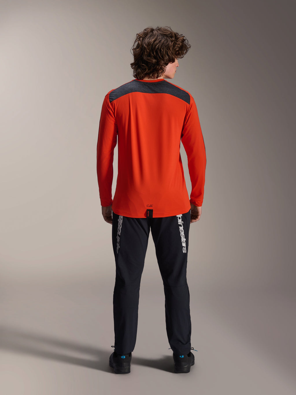Alpinestars A-Dura Insert Astar långärmad tröja i Spicy Orange, vy bakifrån som visar mörkgrå axelpanel, buren med svarta Alpinestars-byxor och svarta skor, professionell studiobild för MTB-utrustning