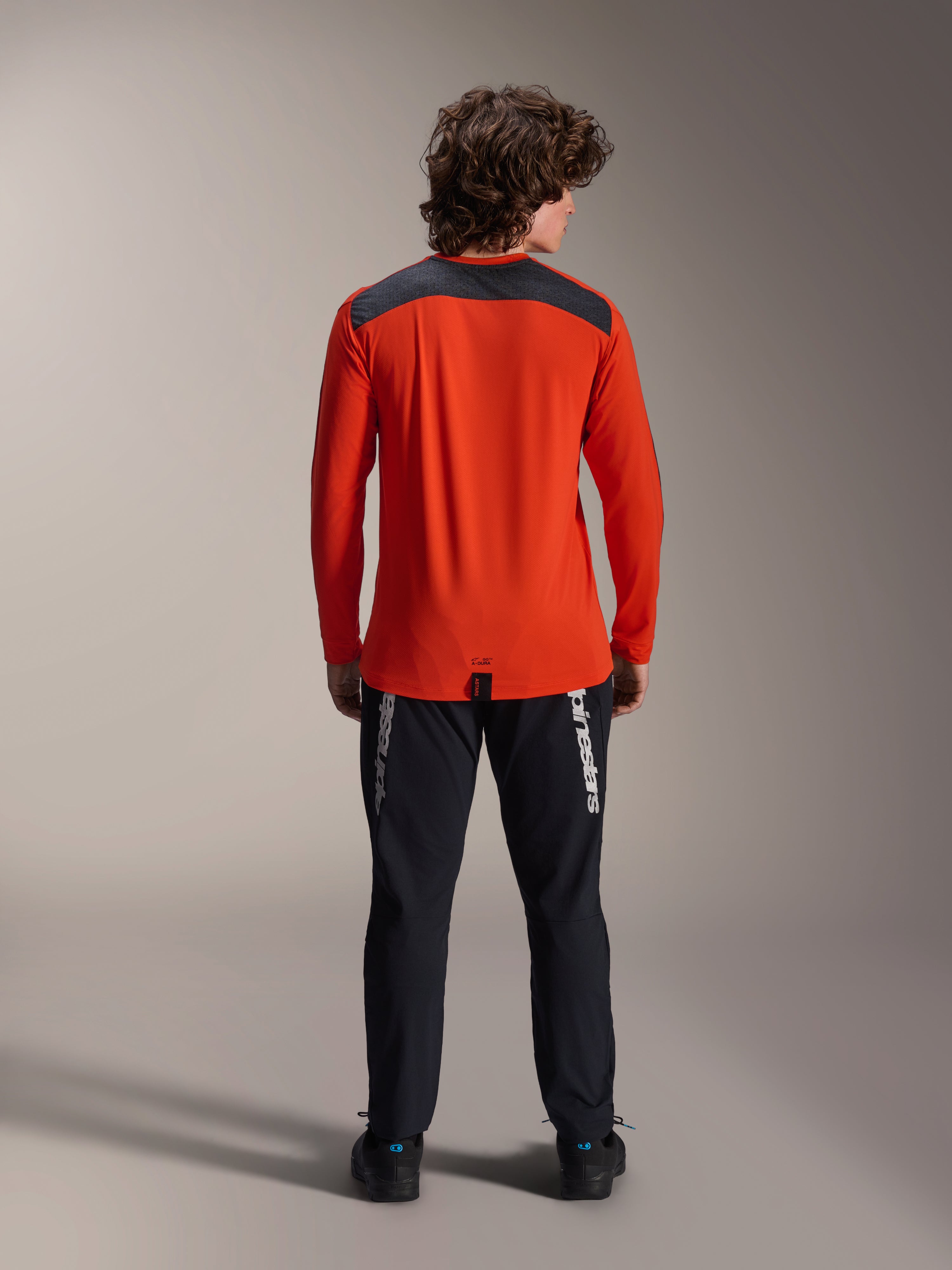 Alpinestars A-Dura Insert Astar långärmad tröja i Spicy Orange, vy bakifrån som visar mörkgrå axelpanel, buren med svarta Alpinestars-byxor och svarta skor, professionell studiobild för MTB-utrustning