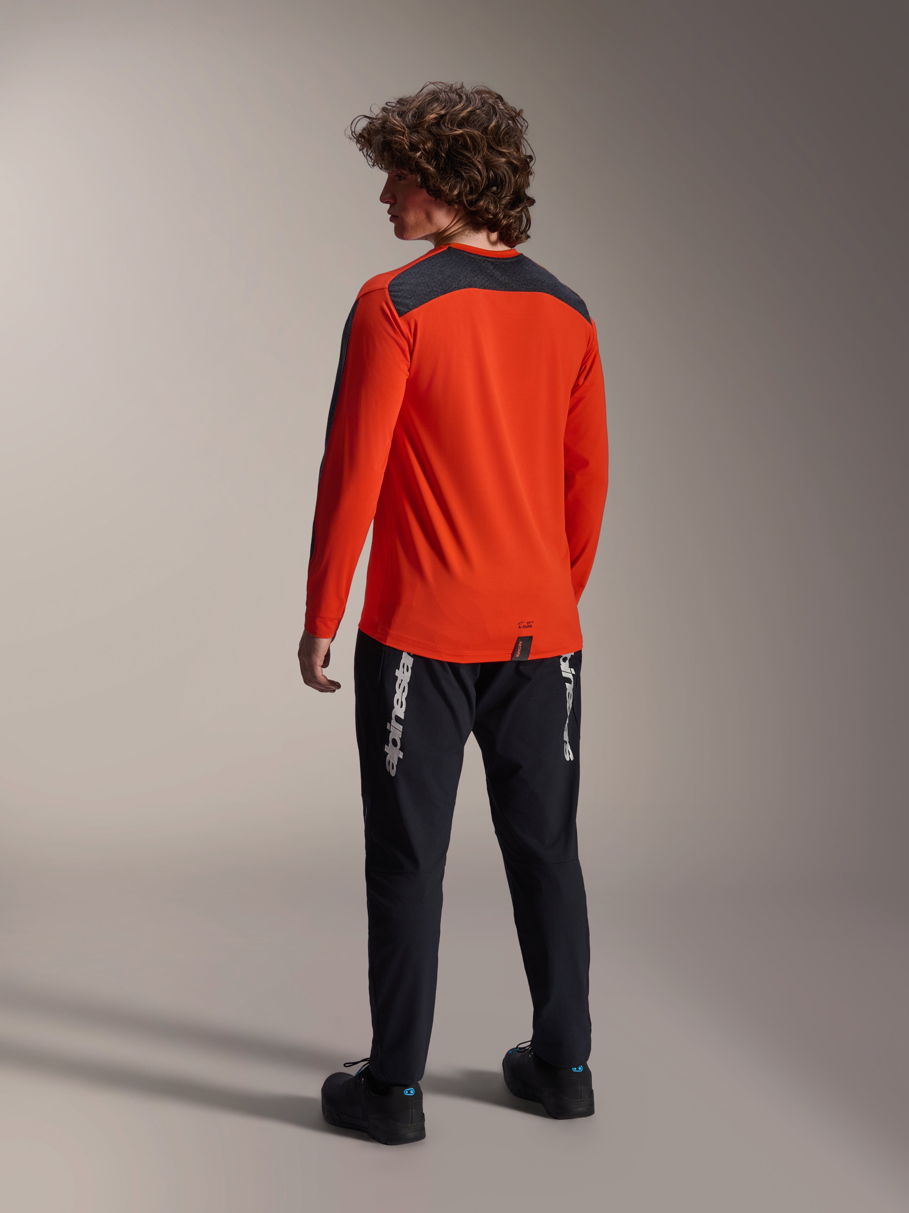 Vy bakifrån av en modell som bär Alpinestars A-Dura Insert Astar-tröjan i Spicy Orange, långärmad MTB-tröja med mörkgrå axelpanel, matchad med svarta märkesbyxor, terrängcyklingsutrustning.