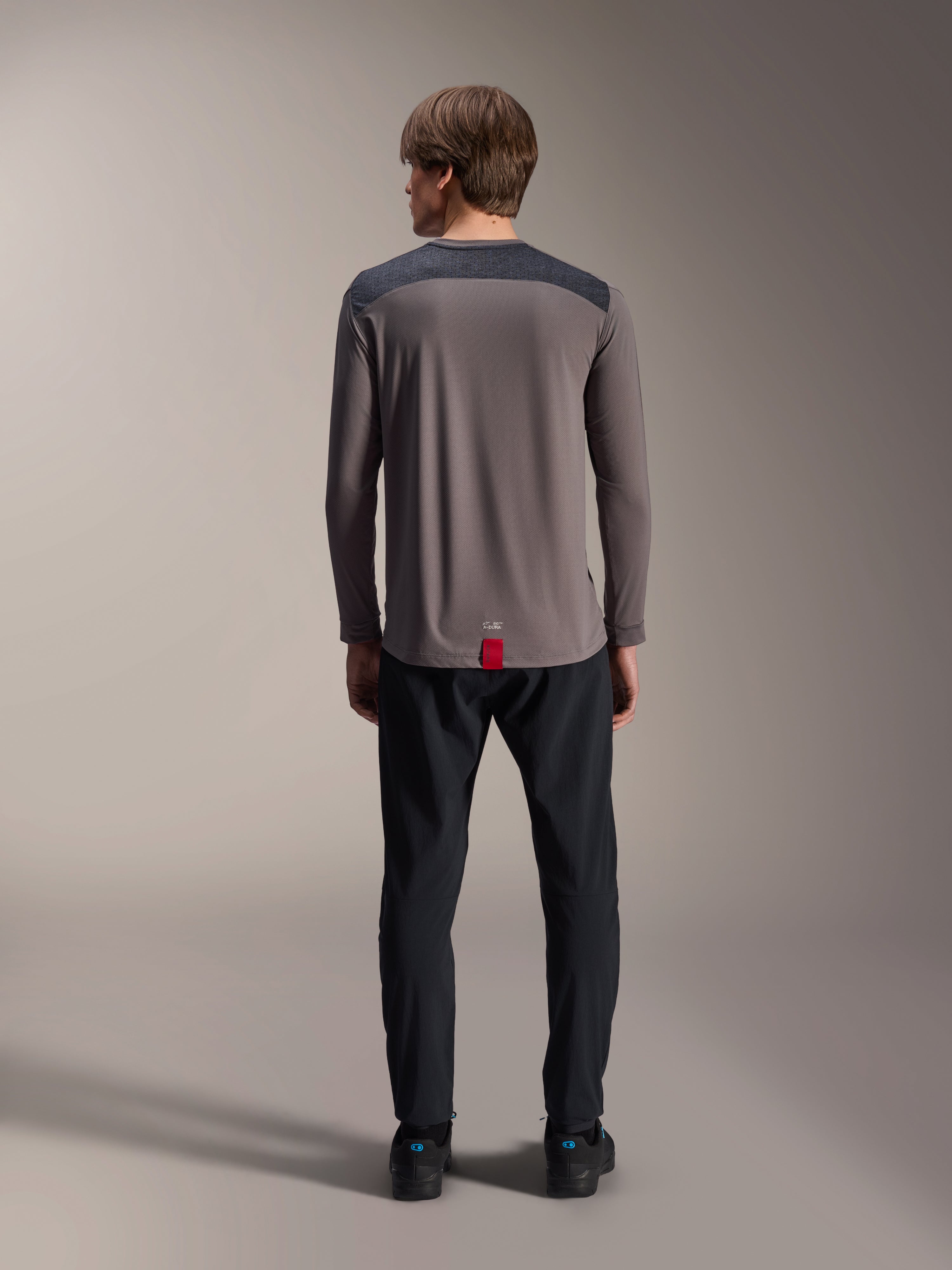 Alpinestars A-Dura Insert Astar långärmad tröja i mörkgrått, vy bakifrån, med koksgrå axelpaneler och en röd flik vid fållen, bärs med svarta byxor och skor, funktionskläder för mountainbike