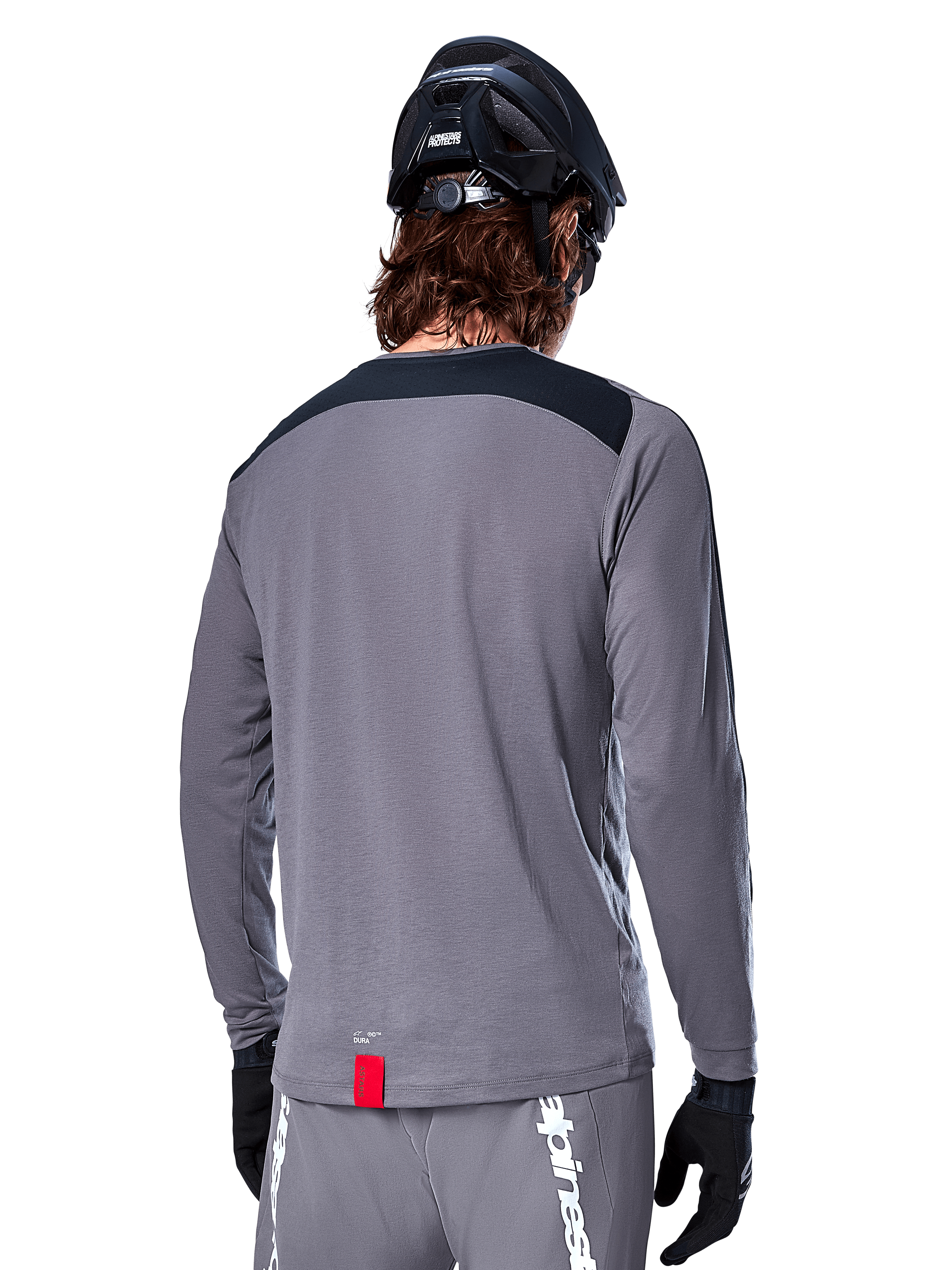 Alpinestars A-Dura Dri Astar långärmad tröja, mörkgrå MTB-tröja sedd bakifrån, med en svart axelpanel och liten röd märkesetikett, buren med en svart mountainbikehjälm och handskar