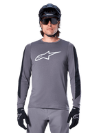Alpinestars A-Dura Dri Astar långärmad tröja, mörkgrå, med en stor vit Astar-logotyp på bröstet och svarta meshpaneler på ärmarna, bärs av en cyklist med en svart mountainbikehjälm och sportglasögon, MTB-cykelkläder