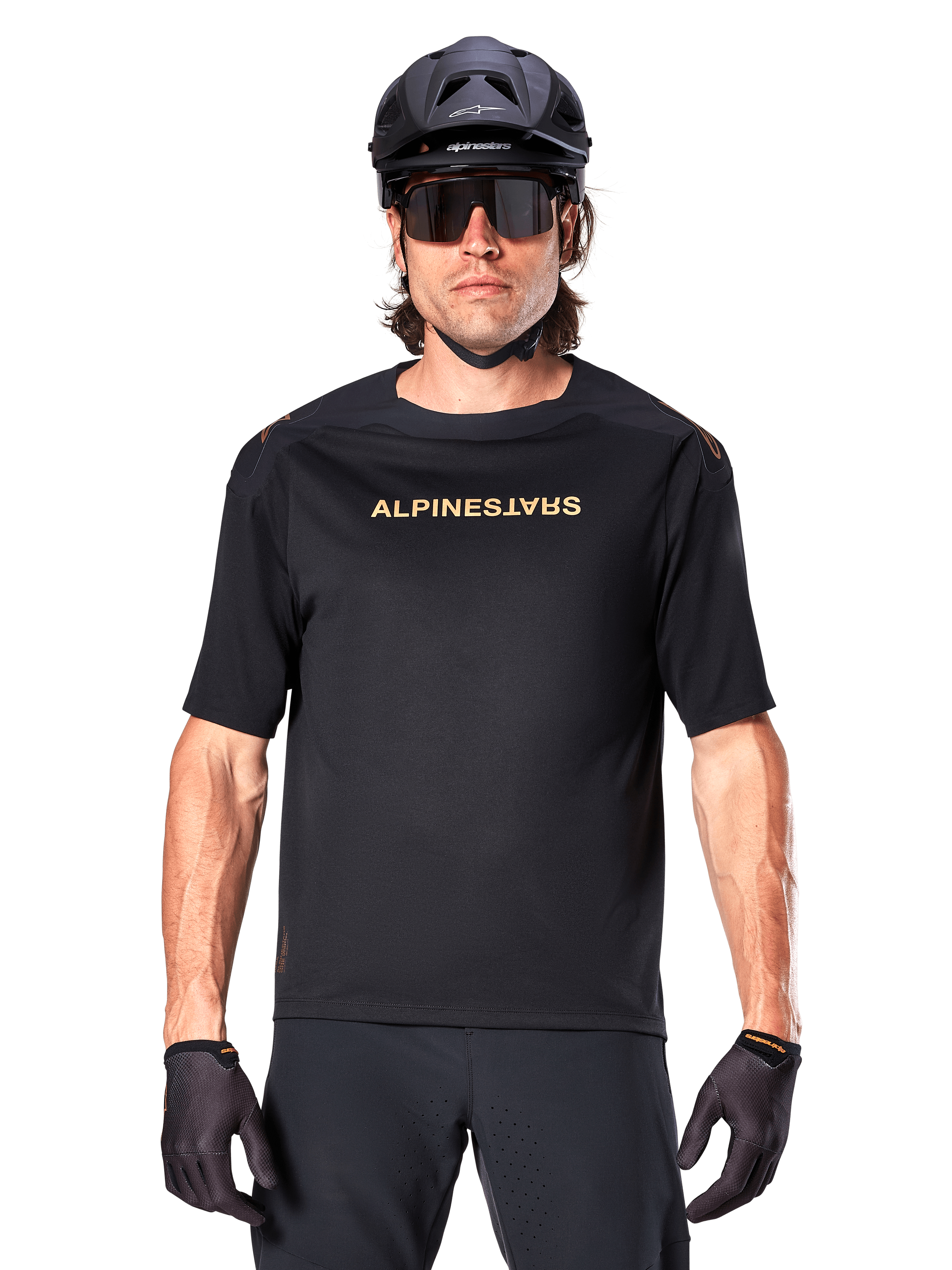 Mountain biker wearing Alpinestars A-Aria Polartec Switch Short Sleeve Jersey in Black, featuring gold-toned chest branding, paired with a black MTB helmet, dark performance sunglasses, and matching gloves, off-road cycling gear.